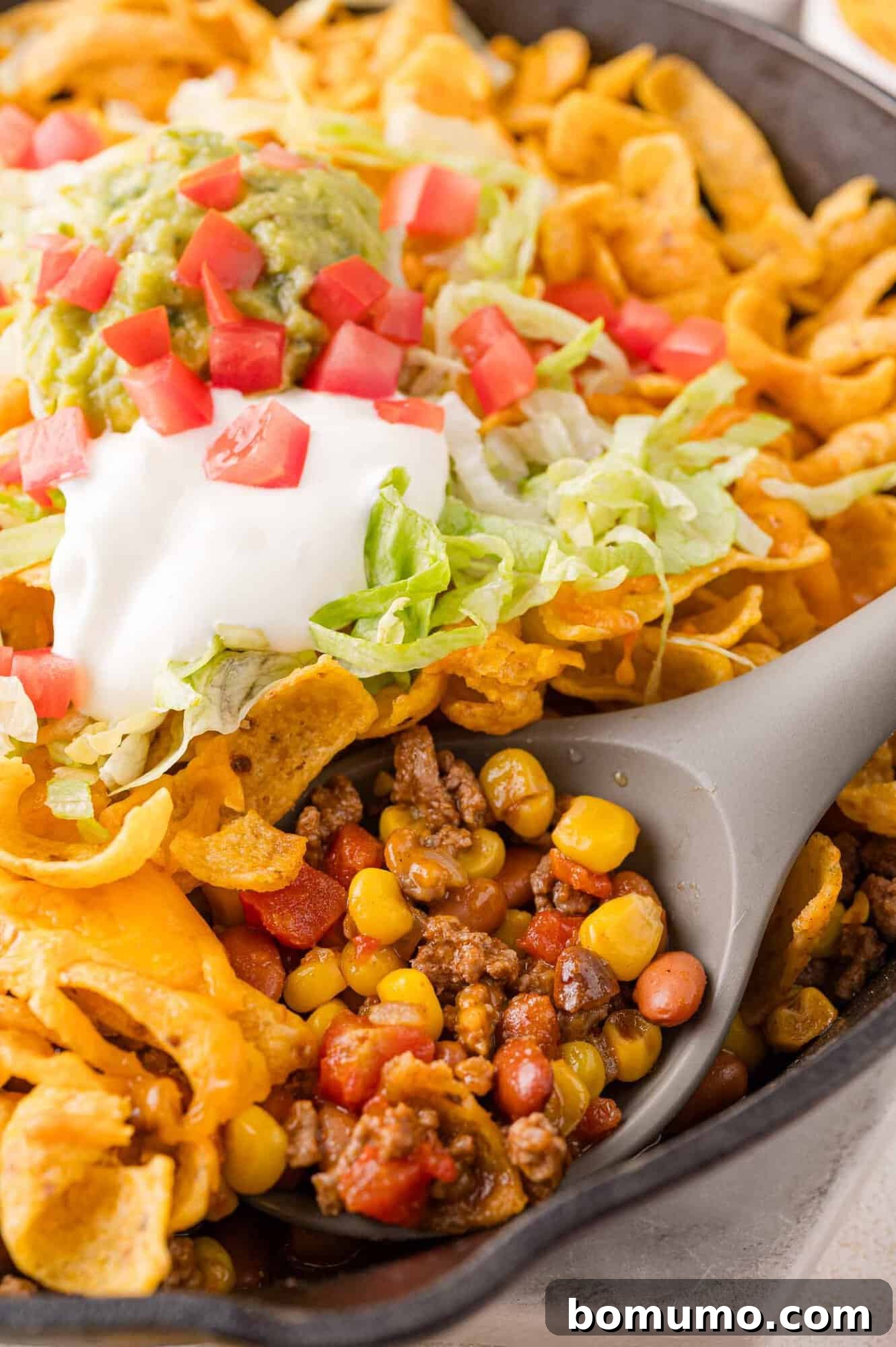 Crunchy Taco Casserole 3 A skillet with a Frito pie, featuring vibrant corn chips, savory ground beef, sweet corn, hearty beans, topped with fresh lettuce, diced tomatoes, cool sour cream, and rich guacamole. A serving spoon is resting in the dish, ready to be enjoyed.