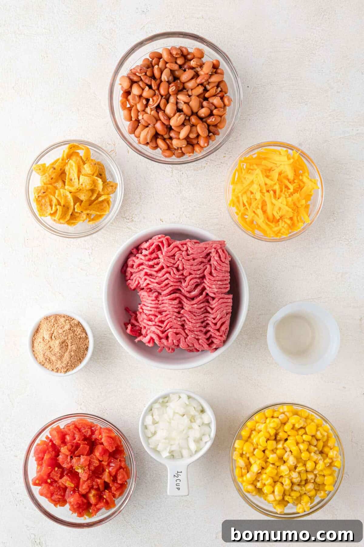 Crunchy Taco Casserole 4 Bowls of ingredients arranged on a surface, showcasing the fresh components for the Walking Taco Casserole: raw ground beef, pinto beans, shredded cheddar cheese, sweet corn, chopped diced tomatoes, crushed corn chips, finely diced onions, a vibrant spice mix, and a small dish of water. Each ingredient is clearly separated, ready for preparation.