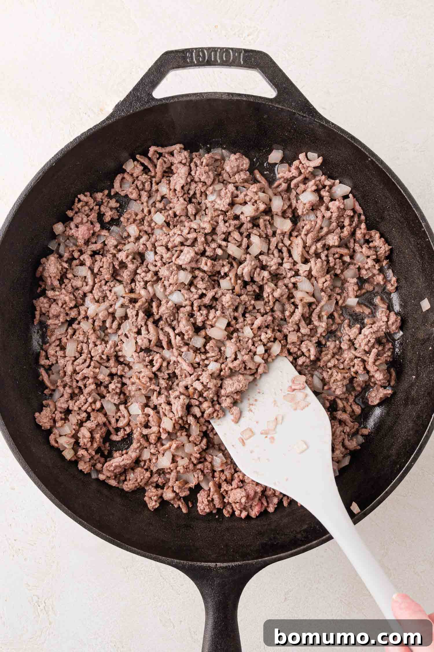 Crunchy Taco Casserole 5 Ground beef and chopped onions being cooked in a black skillet, stirred with a white spatula. The onions are beginning to soften, and the beef is browning, indicating the start of a flavorful base for the casserole.