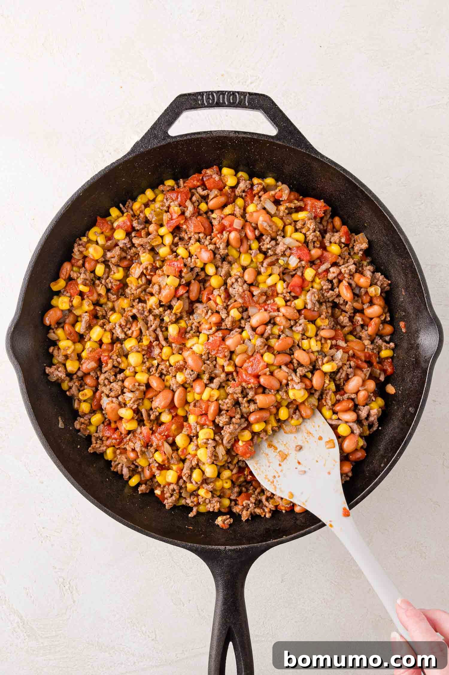 Crunchy Taco Casserole 7 A skillet with a rich mixture of ground beef, pinto beans, corn, and diced tomatoes being stirred with a white spatula. The ingredients are well combined and simmering, preparing to absorb the full flavors of the taco seasoning.
