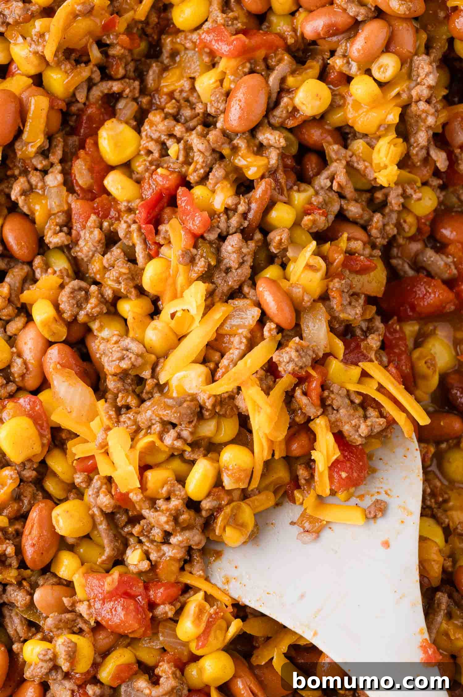 Crunchy Taco Casserole 8 Close-up of a hearty mixture featuring ground beef, sweet corn, kidney beans, vibrant tomatoes, and melted cheese, expertly stirred with a white spatula. The melted cheese creates a creamy, cohesive base for the Walking Taco Casserole.