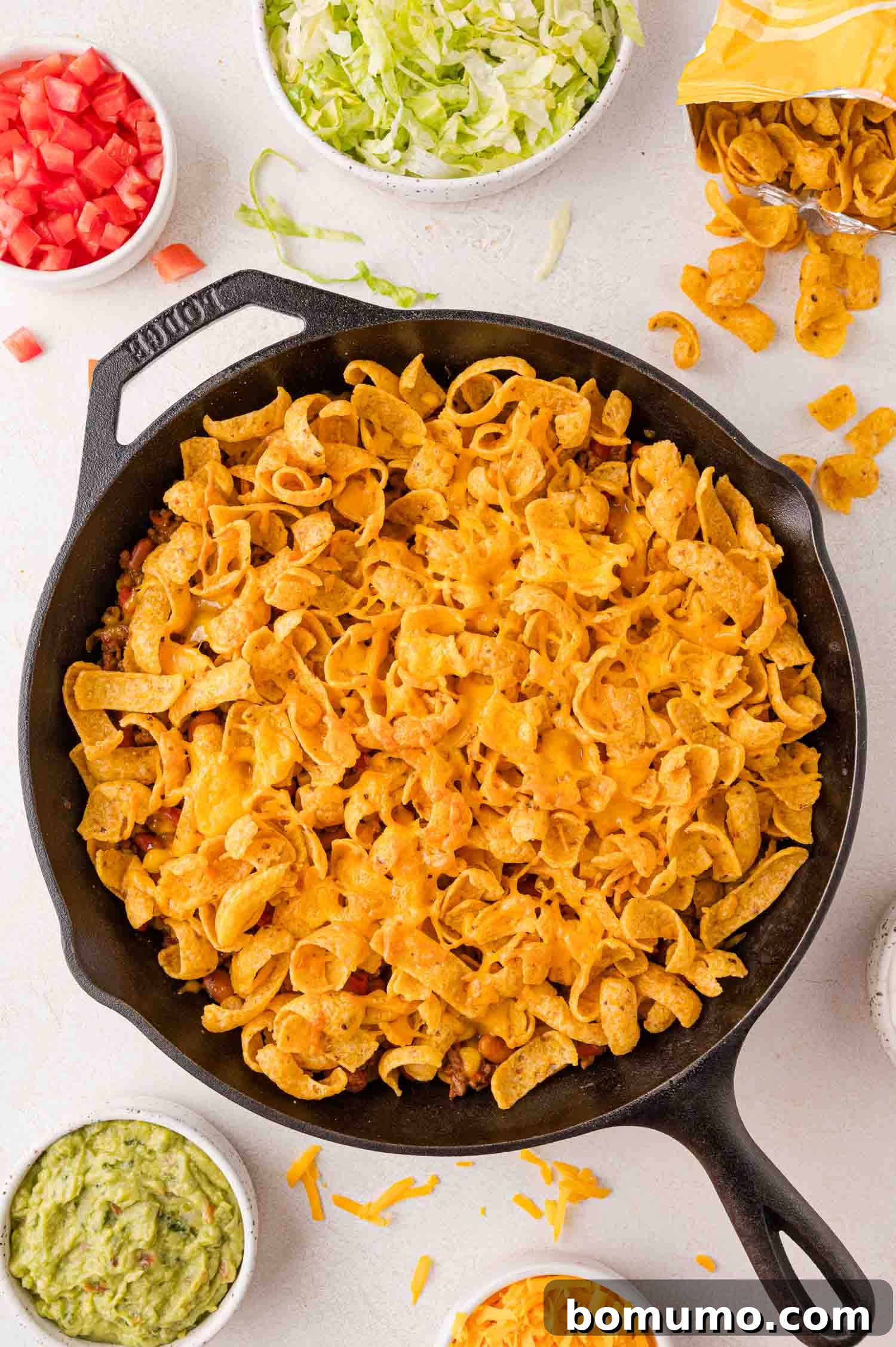 Crunchy Taco Casserole 10 A cast iron skillet filled with a vibrant Walking Taco Casserole, perfectly topped with golden corn chips and deliciously melted cheese. It's surrounded by an inviting array of fresh toppings: bowls of diced tomatoes, crisp shredded lettuce, creamy guacamole, extra shredded cheese, and an open bag of corn chips, ready for customization.