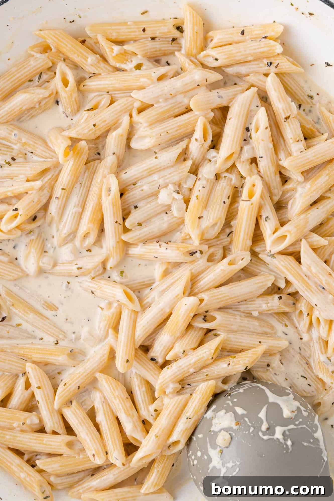 Creamy Chicken Skillet Pasta 12 Penne pasta lovingly coated in a rich, creamy white sauce with vibrant herbs, all stirred in a gleaming skillet with a gray spatula, showing its smooth texture.