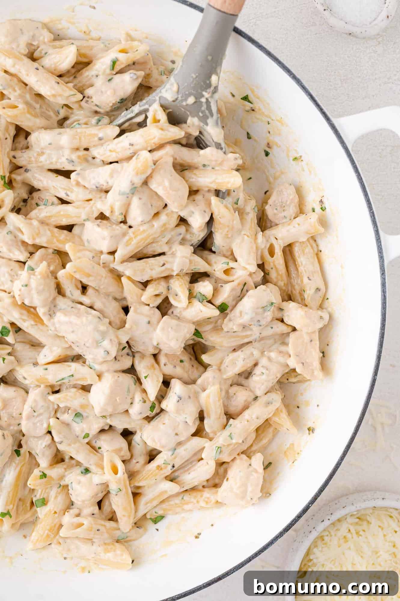 Creamy Chicken Skillet Pasta 16 A bountiful pot of creamy pasta, brimming with succulent pieces of chicken and verdant herbs, being gently stirred with a spoon. A small bowl of grated cheese rests nearby, ready for topping and adding extra flavor.
