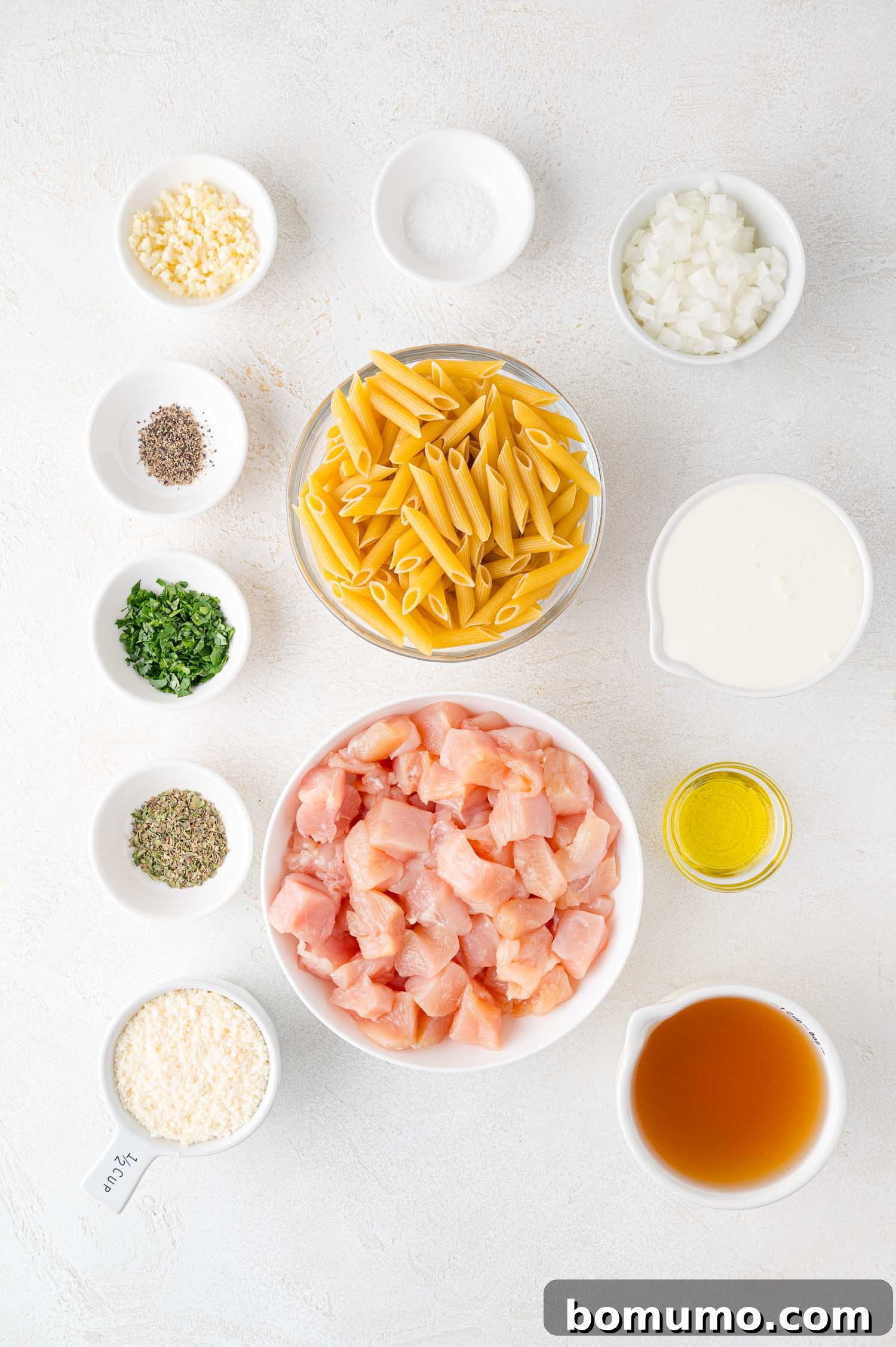 Creamy Chicken Skillet Pasta 4 All the fresh and dry ingredients for a delectable pasta dish are meticulously arranged in individual bowls on a clean white surface. This includes uncooked penne pasta, perfectly diced chicken, finely chopped onions, minced garlic, rich heavy cream, savory chicken broth, golden olive oil, freshly grated Parmesan cheese, and a vibrant array of fresh herbs, ready for cooking.
