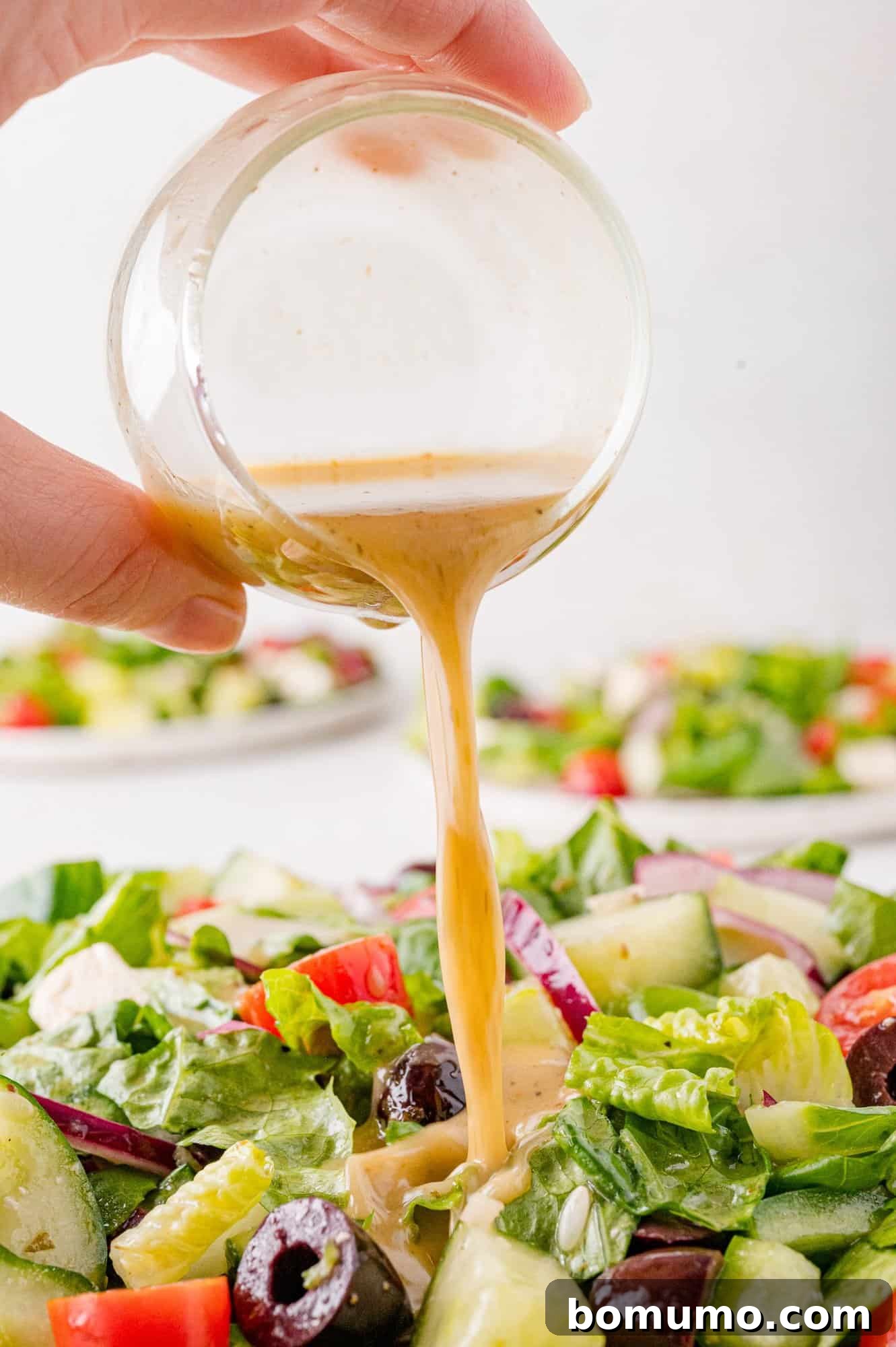 Close-up of a hand pouring Greek salad dressing onto a fresh salad with lettuce, cucumbers, tomatoes, red onions, and olives.