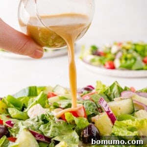 A hand pours dressing over a fresh Greek salad with lettuce, cucumbers, tomatoes, red onions, and olives on a plate.