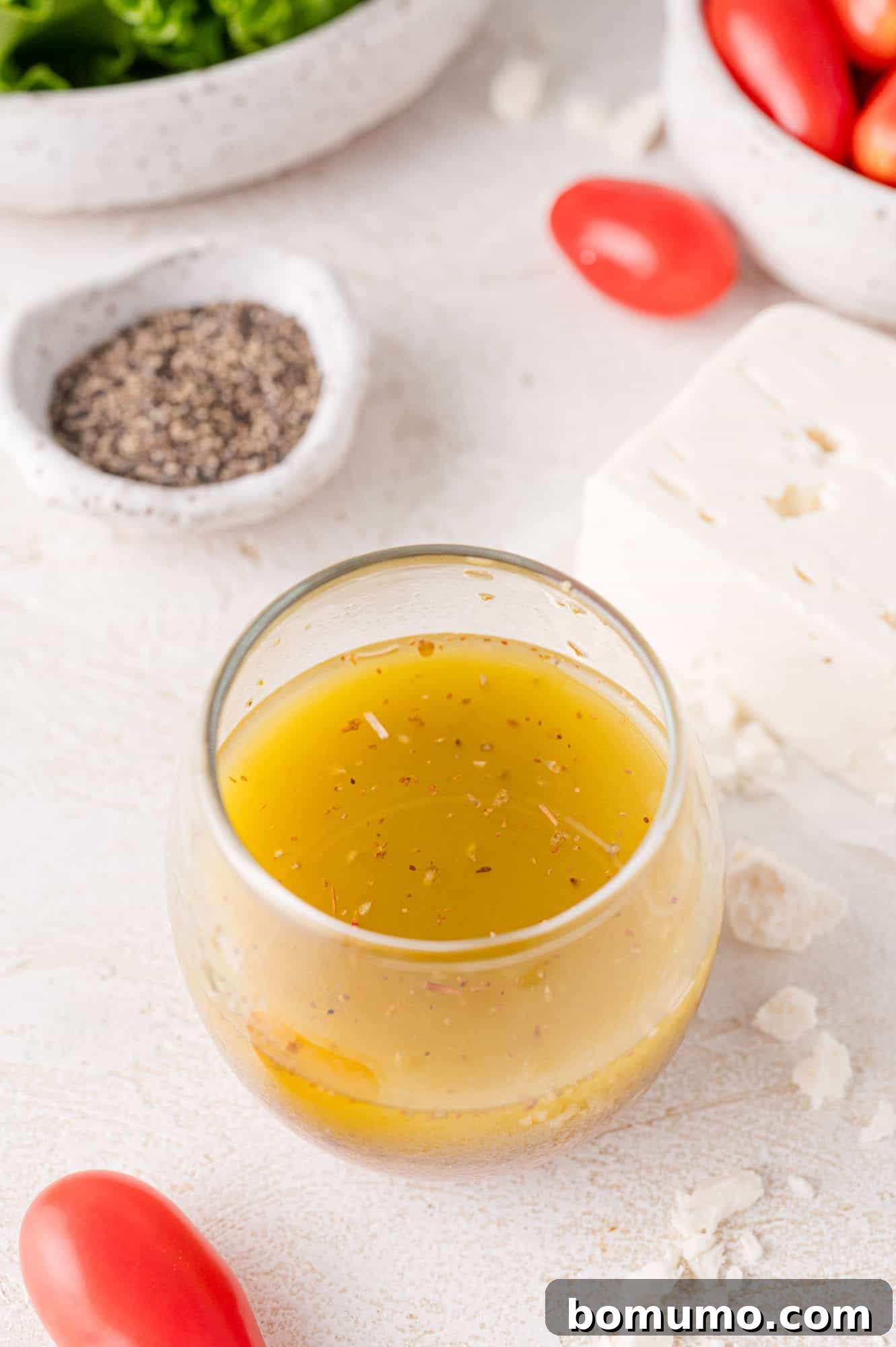 A glass bowl of salad dressing surrounded by pepper, feta cheese, cherry tomatoes, and lettuce on a white surface.