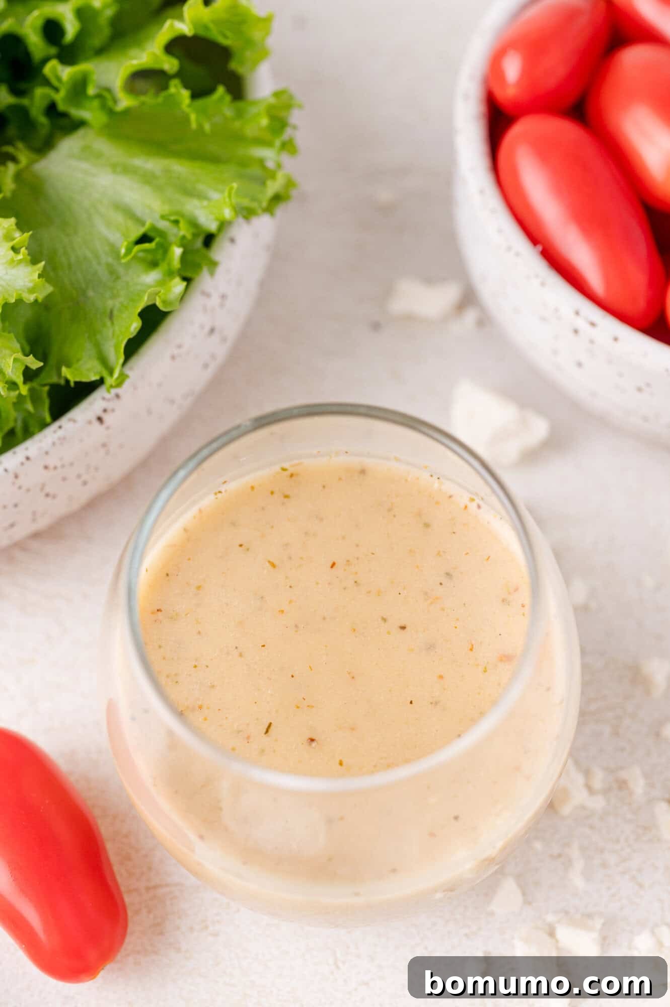 A glass bowl of creamy dressing, surrounded by a bowl of lettuce, cherry tomatoes, and crumbled cheese on a light surface.
