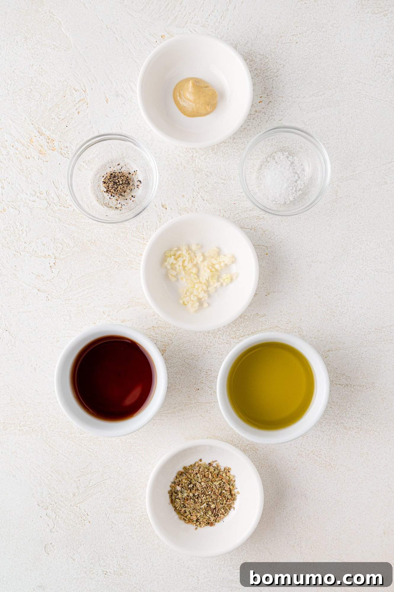 Ingredients arranged on a white surface include olive oil, red vinegar, mustard, minced garlic, dried herbs, salt, and pepper in small white bowls.