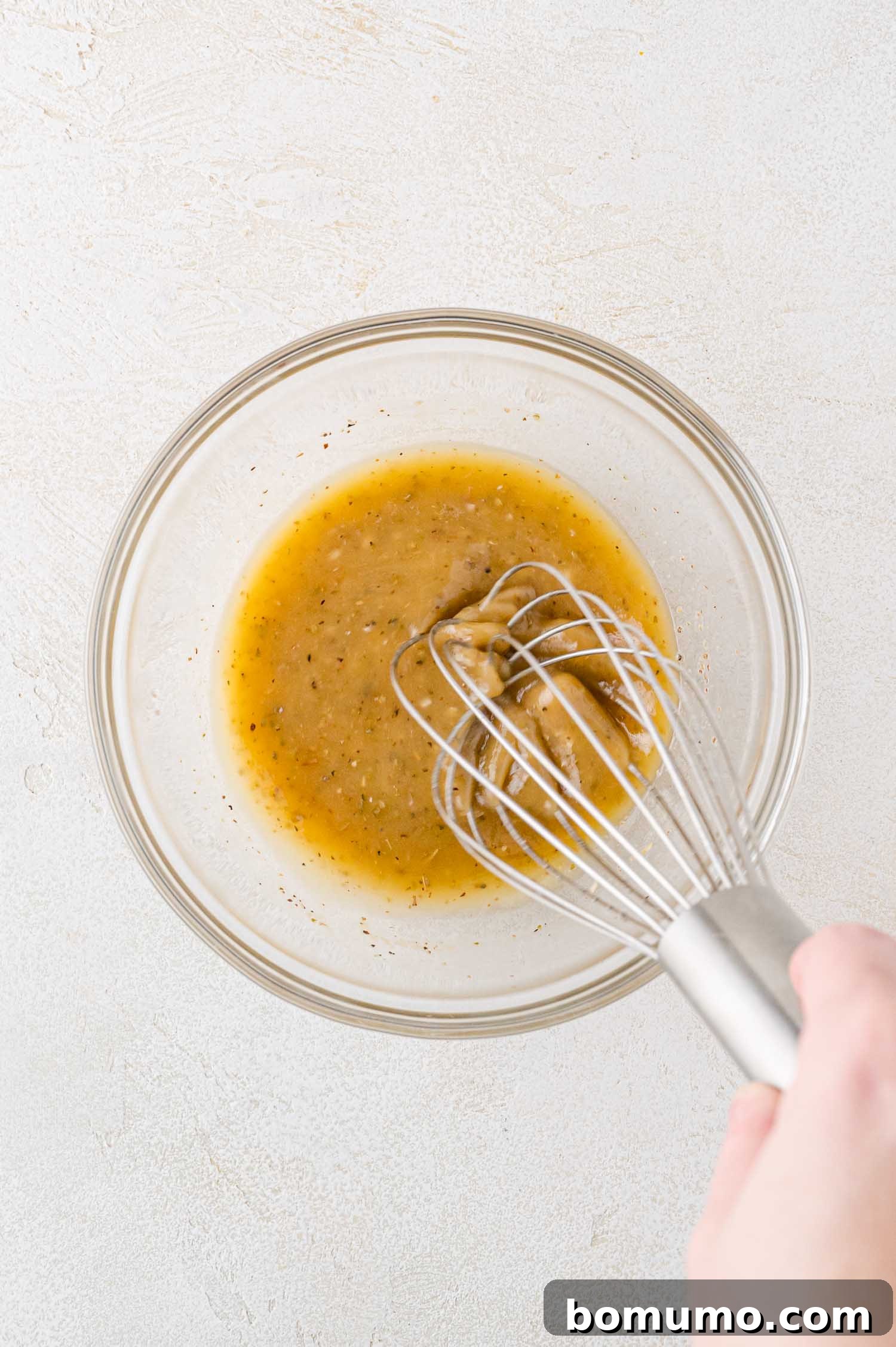 A hand whisking a mixture of oil, vinegar, and herbs in a clear glass bowl on a light textured surface.