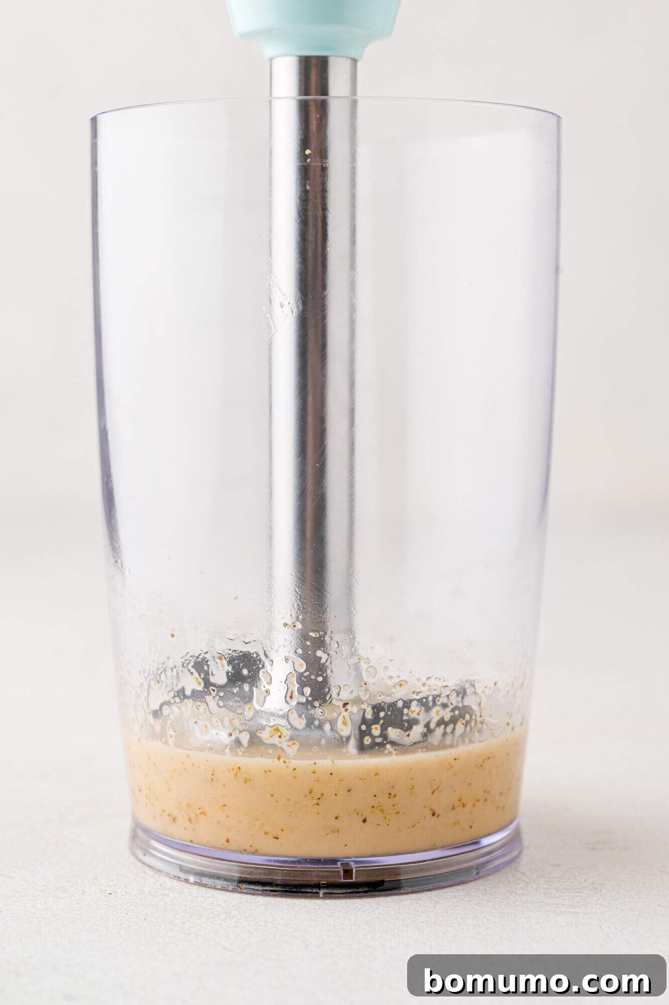 A blender with a metal immersion stick in a clear container, processing a light brown mixture.