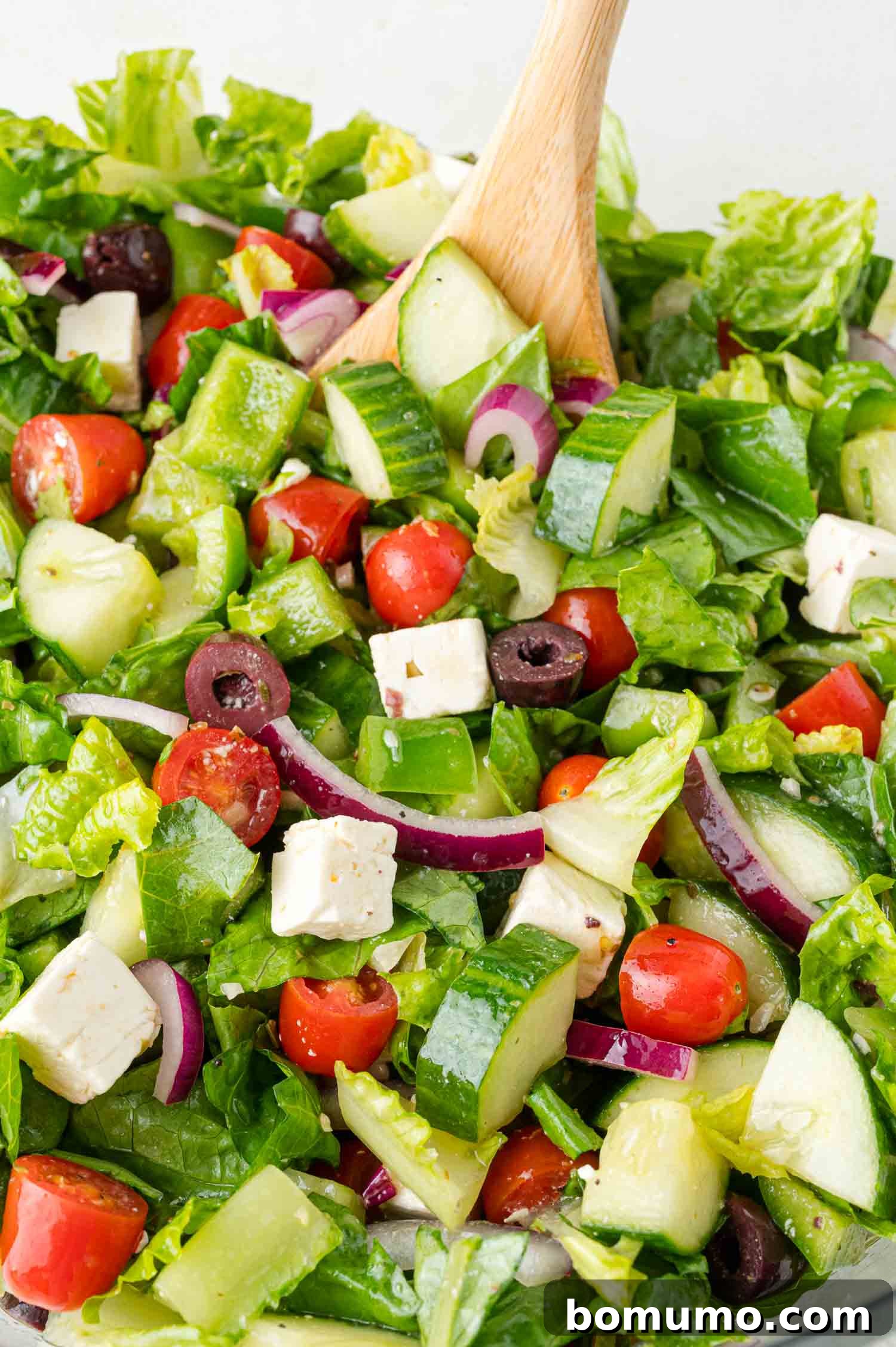 A close-up of a beautifully mixed Greek salad in a clear bowl, showcasing the vibrant colors and fresh ingredients, with a wooden spoon ready for serving.