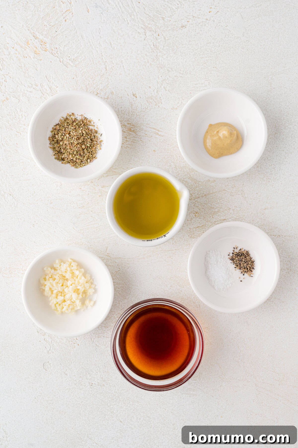 Five small bowls showcasing fresh ingredients for Greek salad dressing, including olive oil, red wine vinegar, oregano, Dijon mustard, salt, pepper, and garlic.