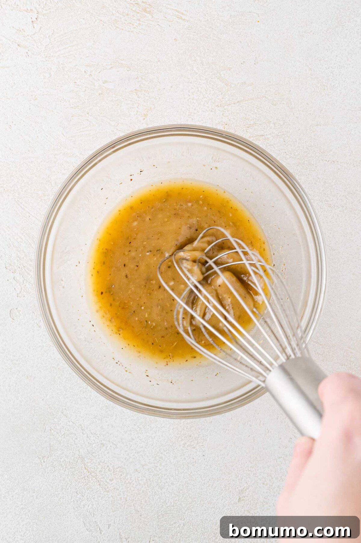 A hand whisking a light golden salad dressing in a clear glass bowl, showing the herbs and seasonings mixing.