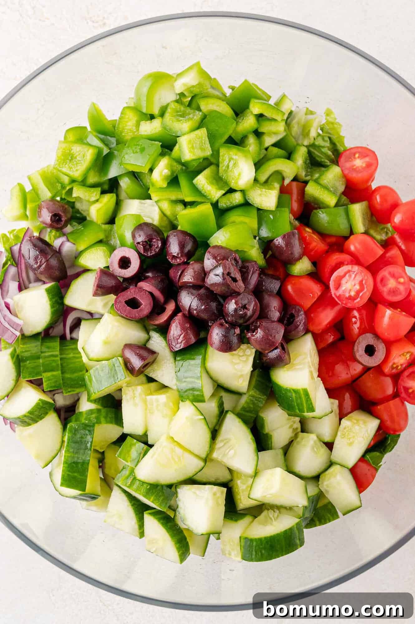 A vibrant assortment of chopped vegetables and olives in a glass bowl, including cucumbers, tomatoes, green bell peppers, red onions, and Kalamata olives, all prepped for Greek salad.