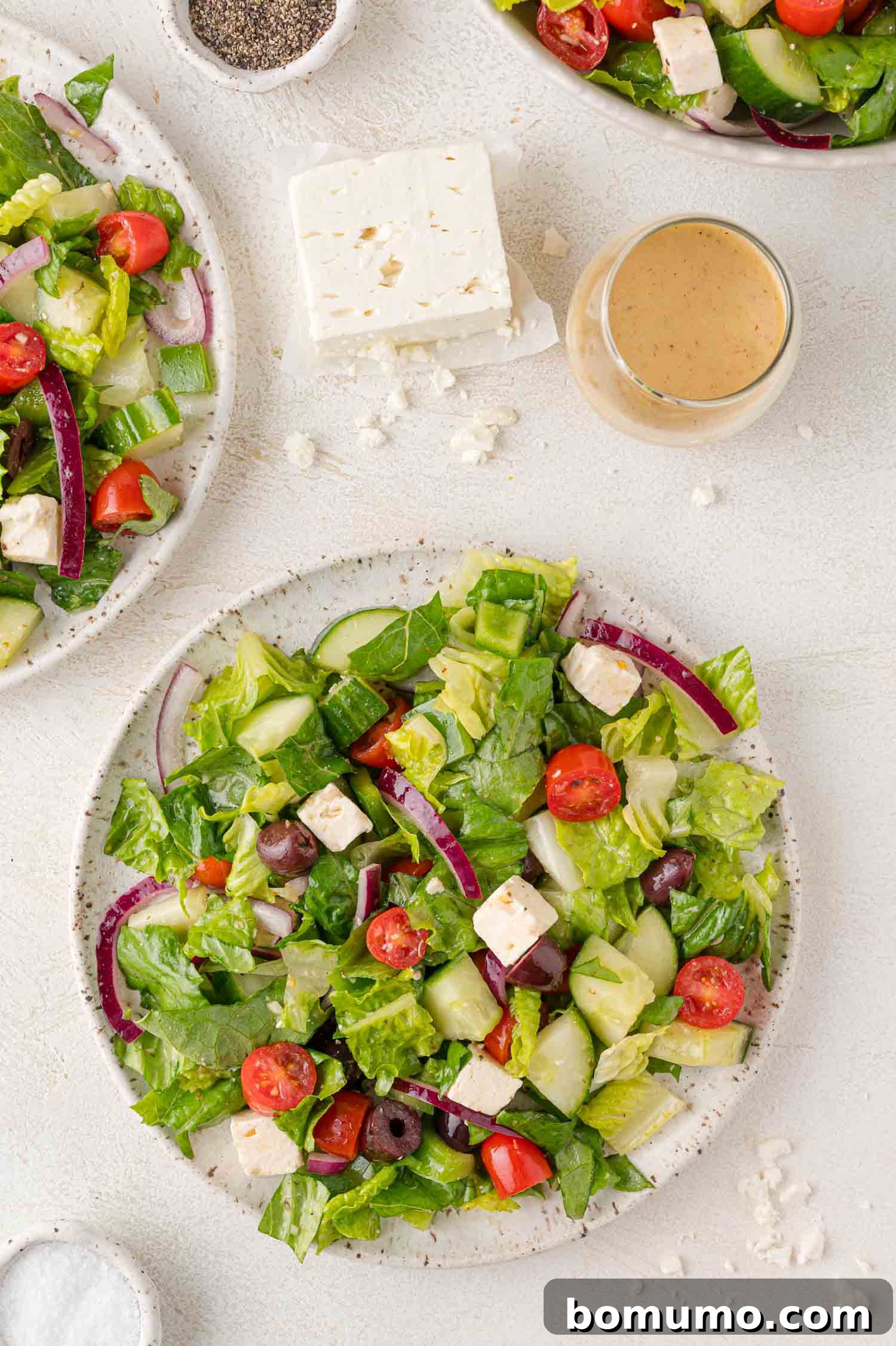 A beautifully presented plate of Greek salad with crisp lettuce, colorful tomatoes, cucumbers, red onion, olives, and feta cheese, served alongside a glass of dressing and seasoning.