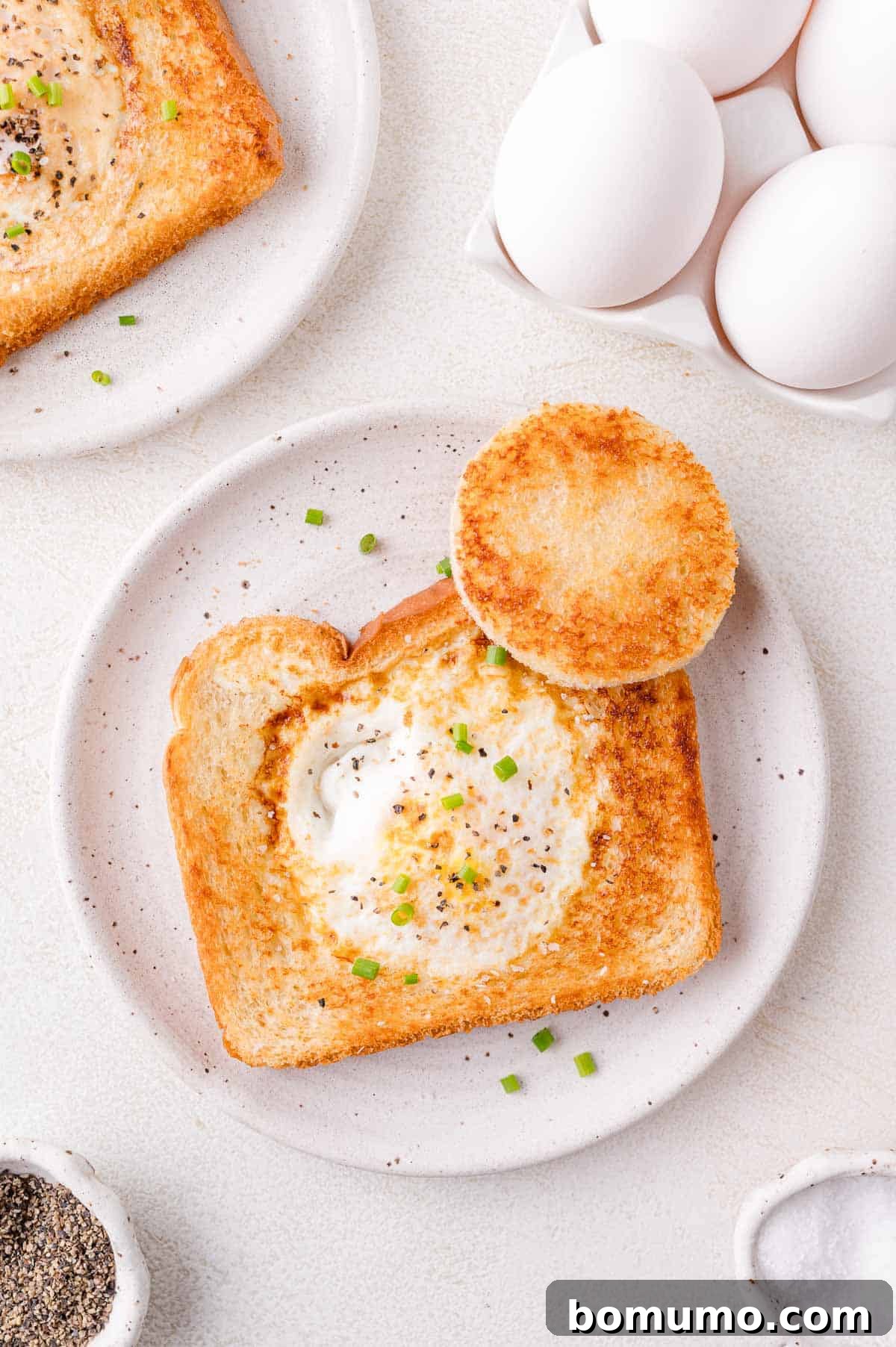 Toasted bread with a circular cutout holds a cooked egg, garnished with chopped chives, accompanied by a round bread piece on a white plate. Nearby are eggs in a carton and seasoning bowls.
