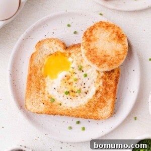 A piece of toast with a cooked egg in the center, garnished with chopped chives, served on a white plate with a toasted bread circle on the side.