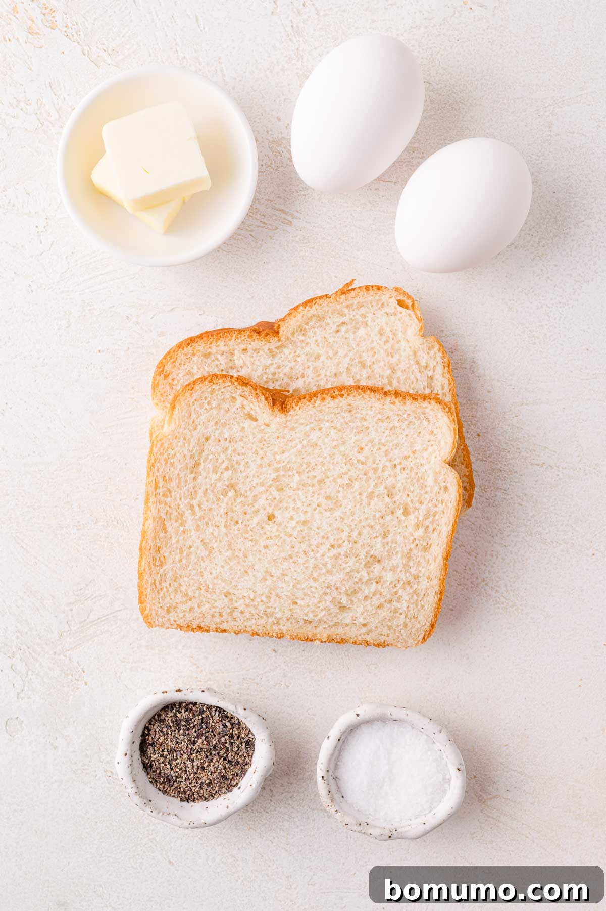 Two slices of bread, two eggs, a small dish of butter, a bowl of salt, and a bowl of pepper on a light surface.