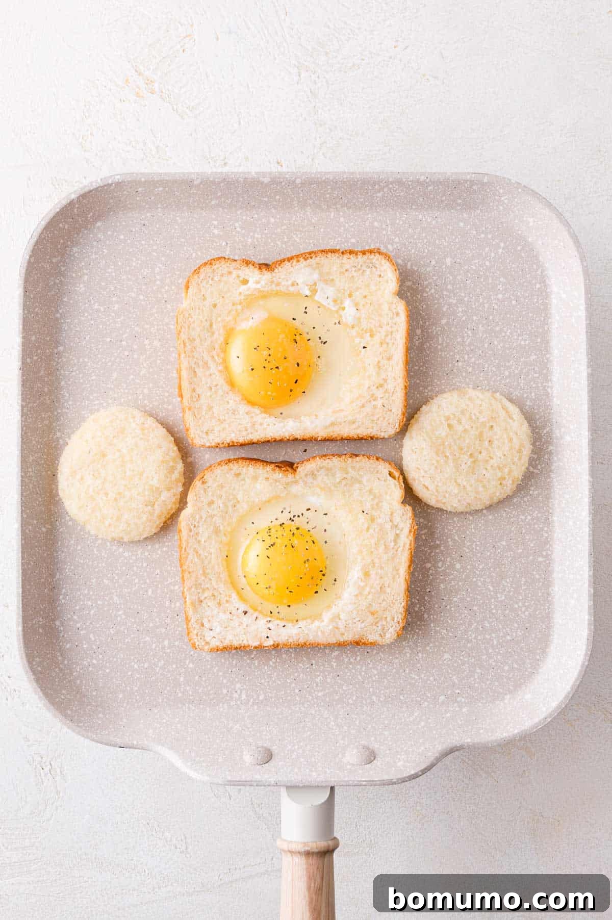 Two slices of bread each with a raw egg in the center are on a frying pan. Two round cutouts of bread are placed next to them.