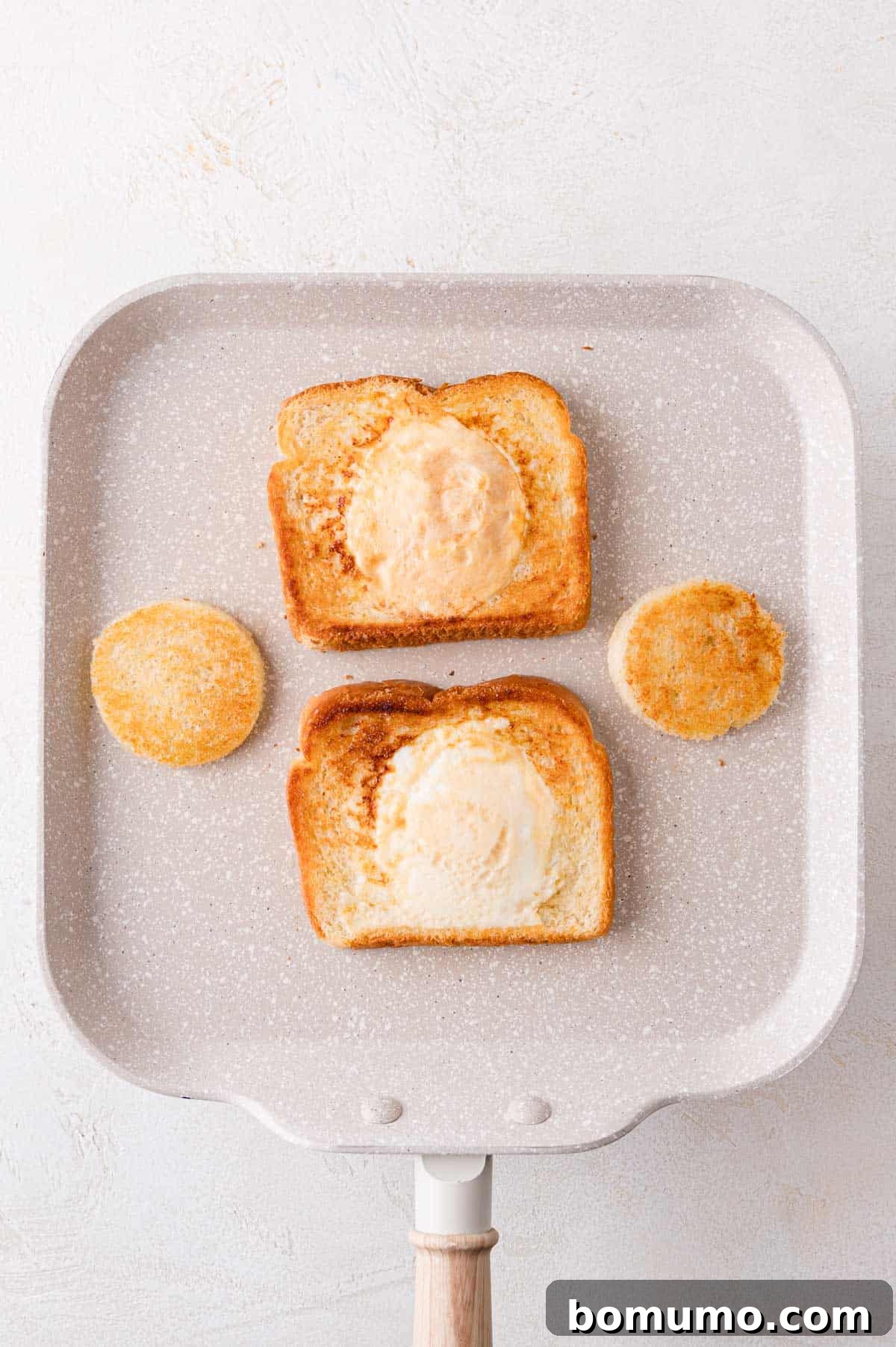 Two slices of toasted bread with circular cutouts are on a square pan. Fried eggs fill the cutouts. Two toasted bread circles are next to the slices.