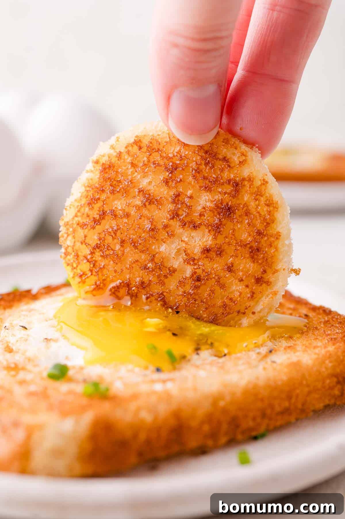 Hand dipping a round, toasted bread piece into a runny egg yolk on a slice of bread.