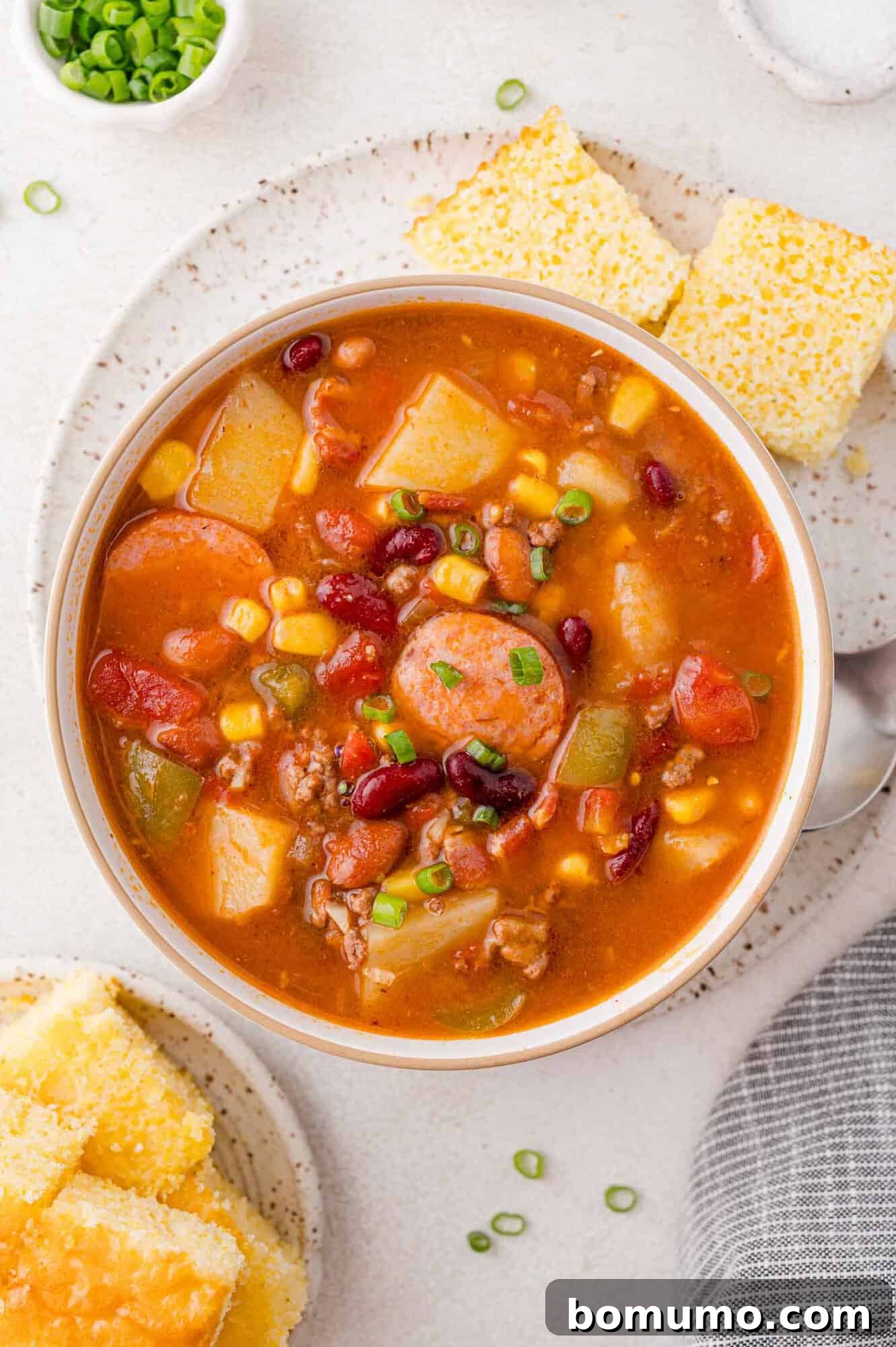 A vibrant bowl of cowboy stew brimming with corn, diced tomatoes, assorted beans, and bell peppers, elegantly garnished with fresh green onions. Alongside, perfectly golden cornbread squares await.