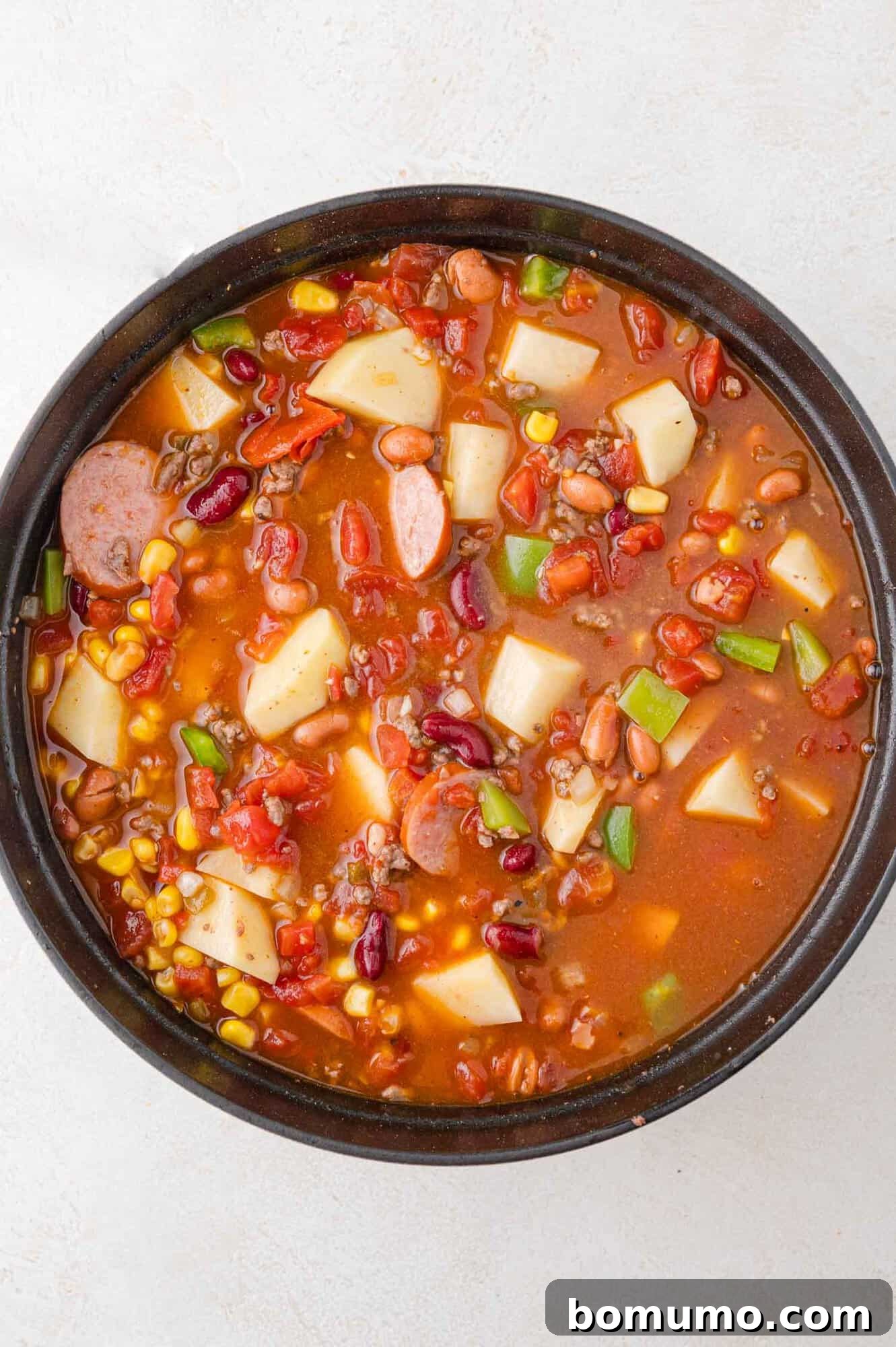 A robust bowl of hearty stew, filled with savory sausage, tender potatoes, a mix of beans, sweet corn, juicy tomatoes, and vibrant green bell peppers, all bathed in a rich red broth.