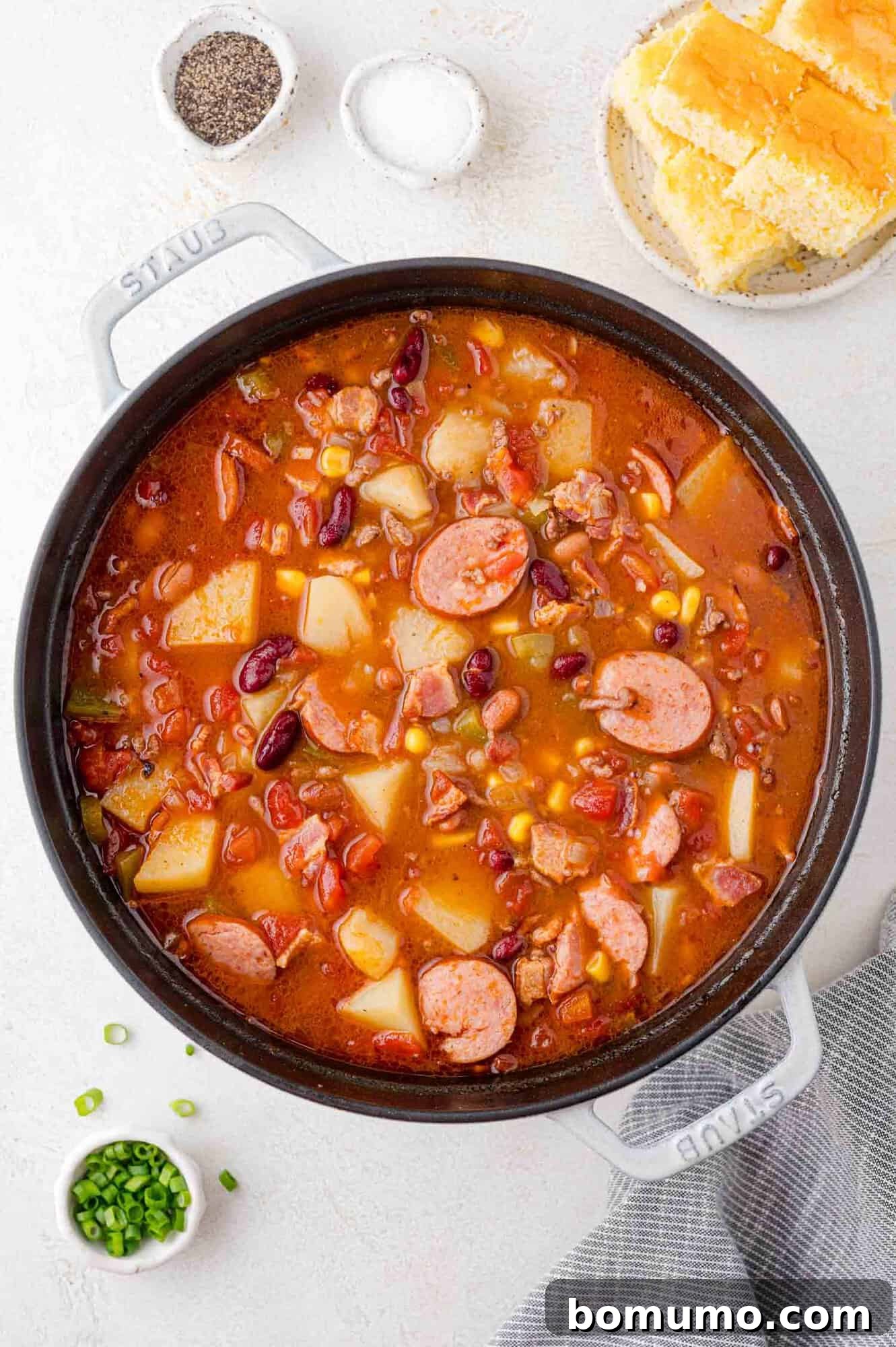 A pot of deliciously cooked stew, brimming with savory sausages, tender potatoes, a colorful mix of beans and vegetables. Small bowls of pepper, chopped green onions, and a plate with golden cornbread are thoughtfully arranged on the side.