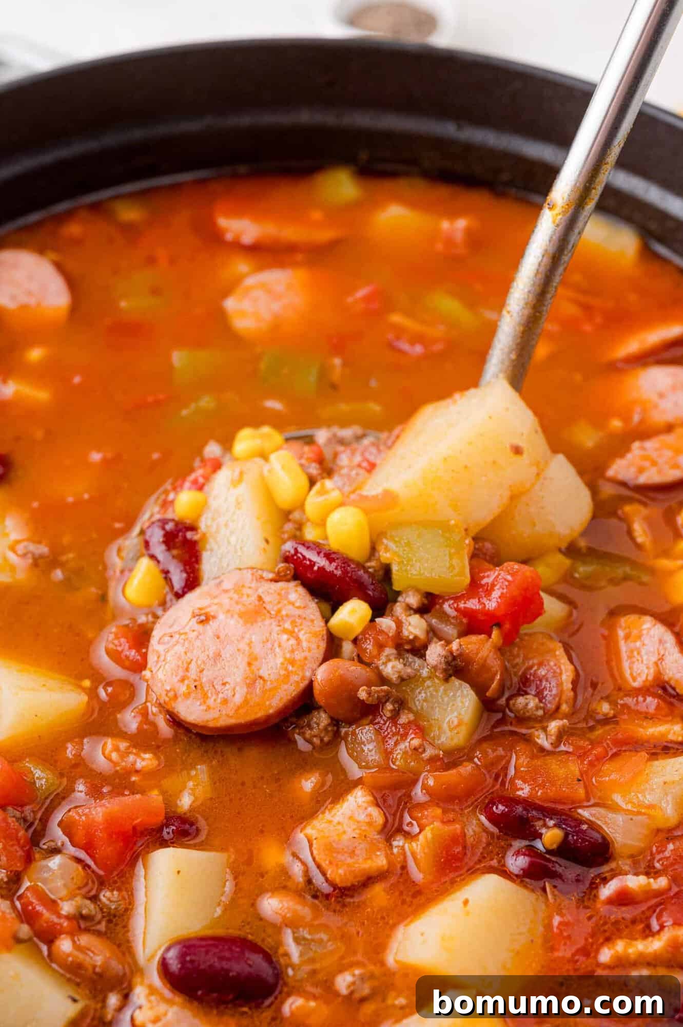 Close-up of a pot filled with appetizing stew, prominently featuring savory sliced sausage, tender potatoes, a mix of beans, sweet corn, and rich tomatoes, with a gleaming metal ladle poised to scoop a perfect portion.
