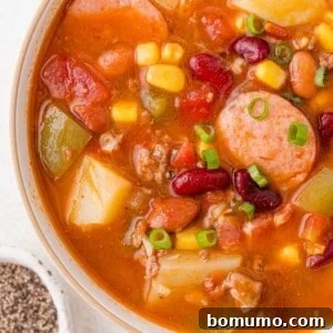 A bowl of hearty stew with sausage slices, beans, corn, potatoes, and diced vegetables garnished with chopped green onions.