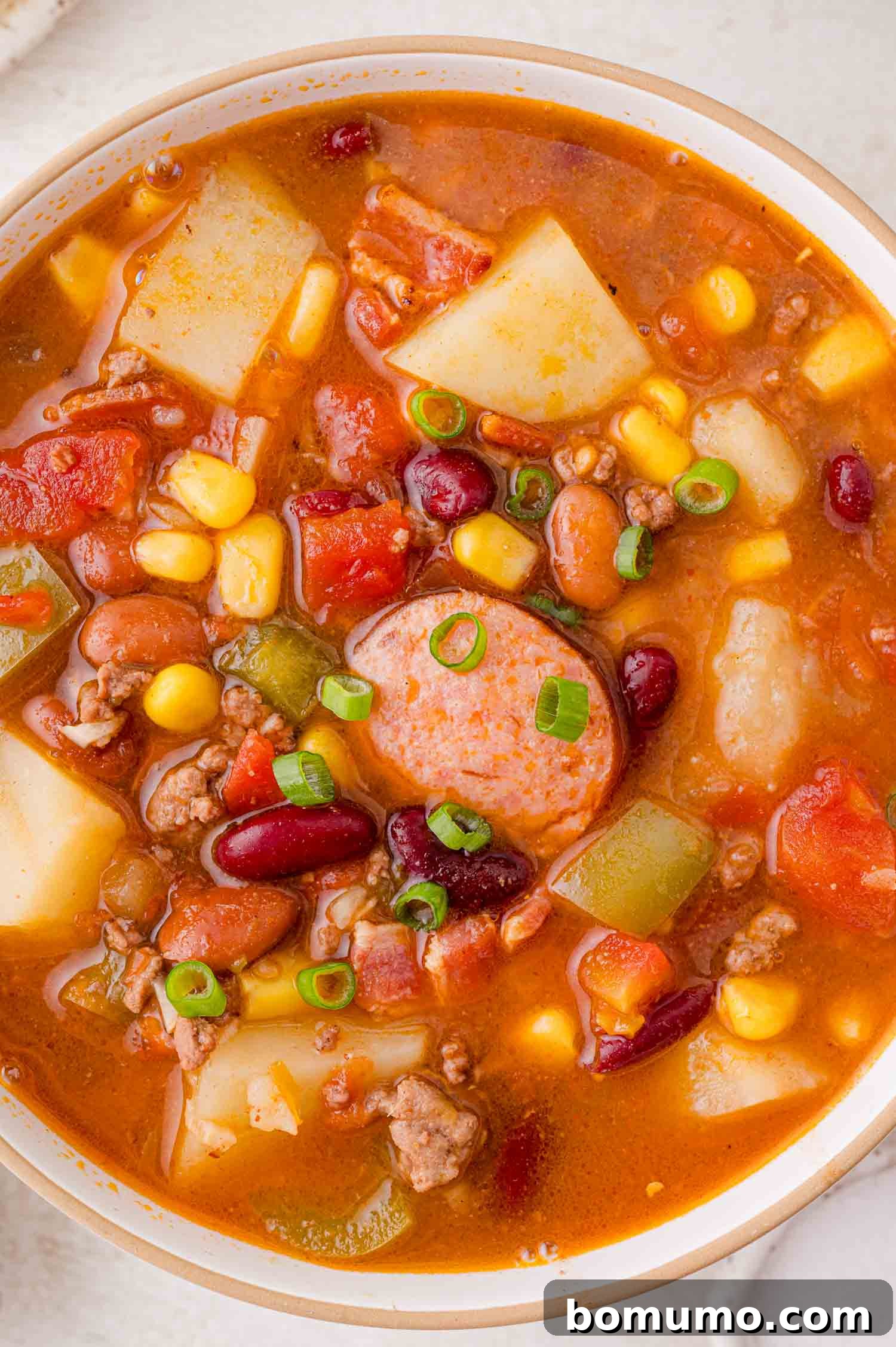 A generously filled bowl of hearty cowboy stew, showcasing savory sausage, tender potatoes, a mix of beans, golden corn, vibrant green peppers, and rich diced tomatoes, all crowned with a sprinkle of sliced green onions.