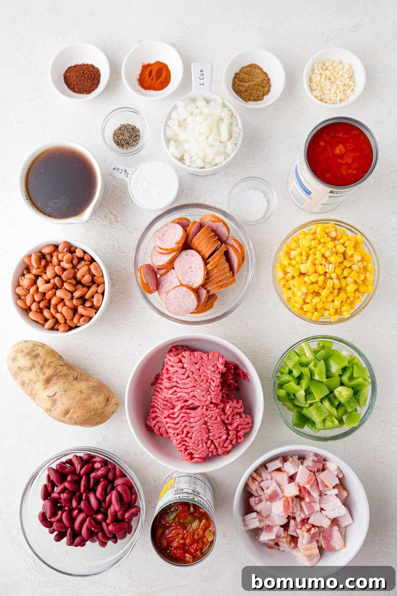 An assortment of fresh and canned ingredients meticulously arranged on a clean white surface, showcasing the components of a robust cowboy stew. Visible are various beans, raw ground meat, strips of bacon, vibrant corn, a bright green bell pepper, savory sausages, a russet potato, canned tomatoes, and a collection of different seasonings in small bowls.