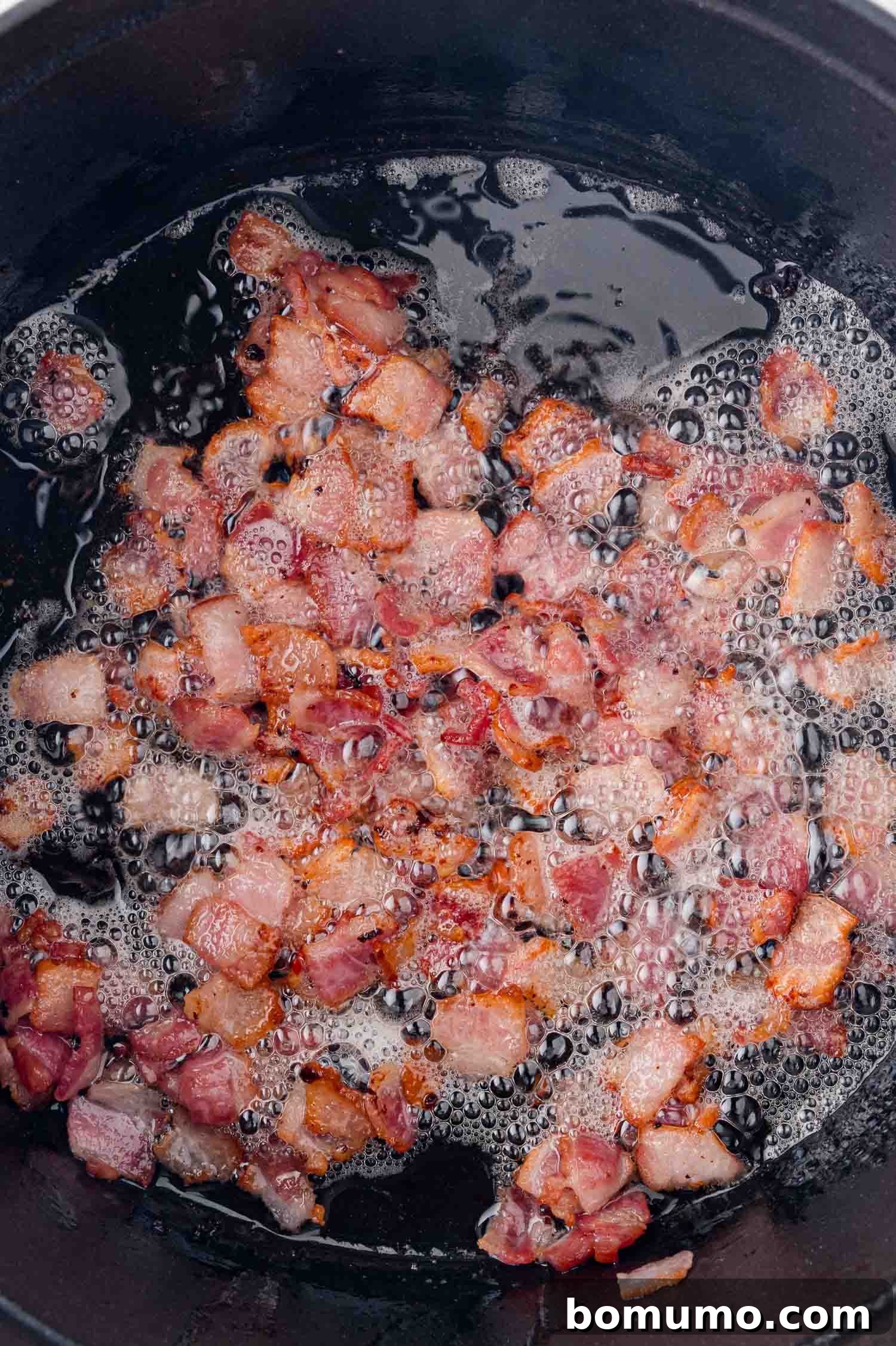 Close-up of diced bacon frying in a black skillet, surrounded by bubbling oil, creating a rich aroma and crispy texture.