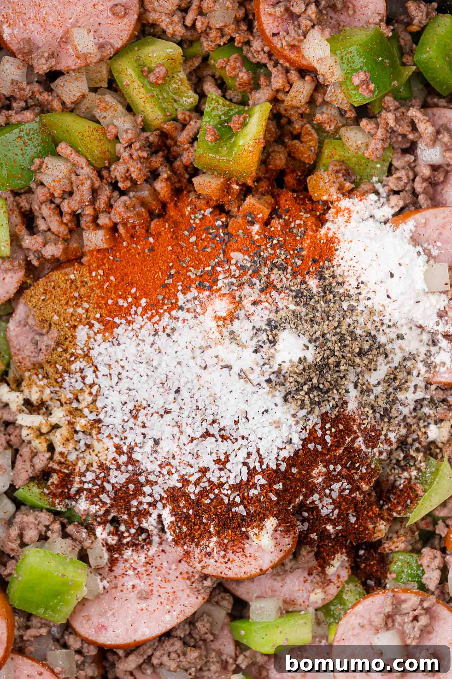 Close-up of chopped green bell peppers, ground beef, sliced sausage, diced onions, and a colorful mix of spices including salt, pepper, and paprika, awaiting a thorough mixing.