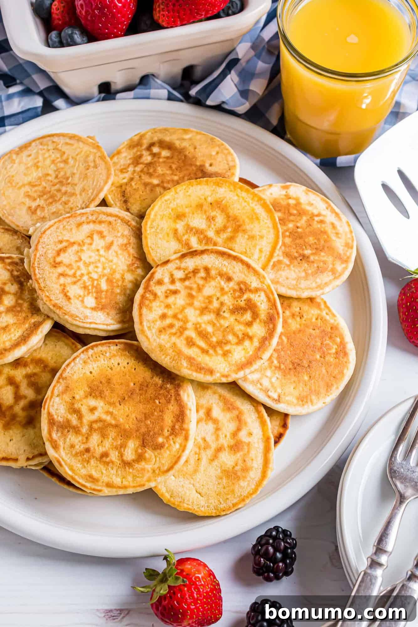 A plate of golden silver dollar pancakes surrounded by strawberries, blackberries, and a glass of orange juice on a table with a blue checkered cloth.