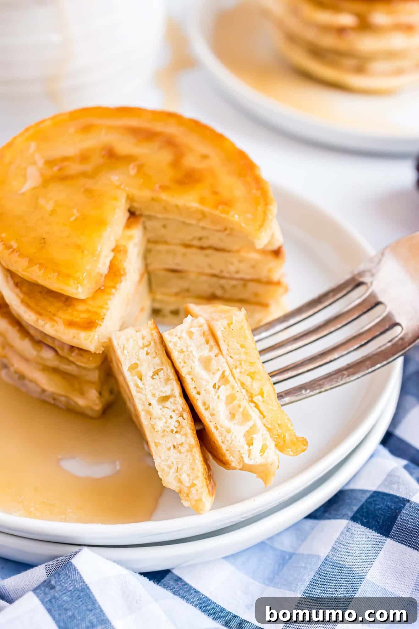 A fork holding a section of a pancake stack with syrup drizzled over, served on a white plate, with a checkered cloth underneath.