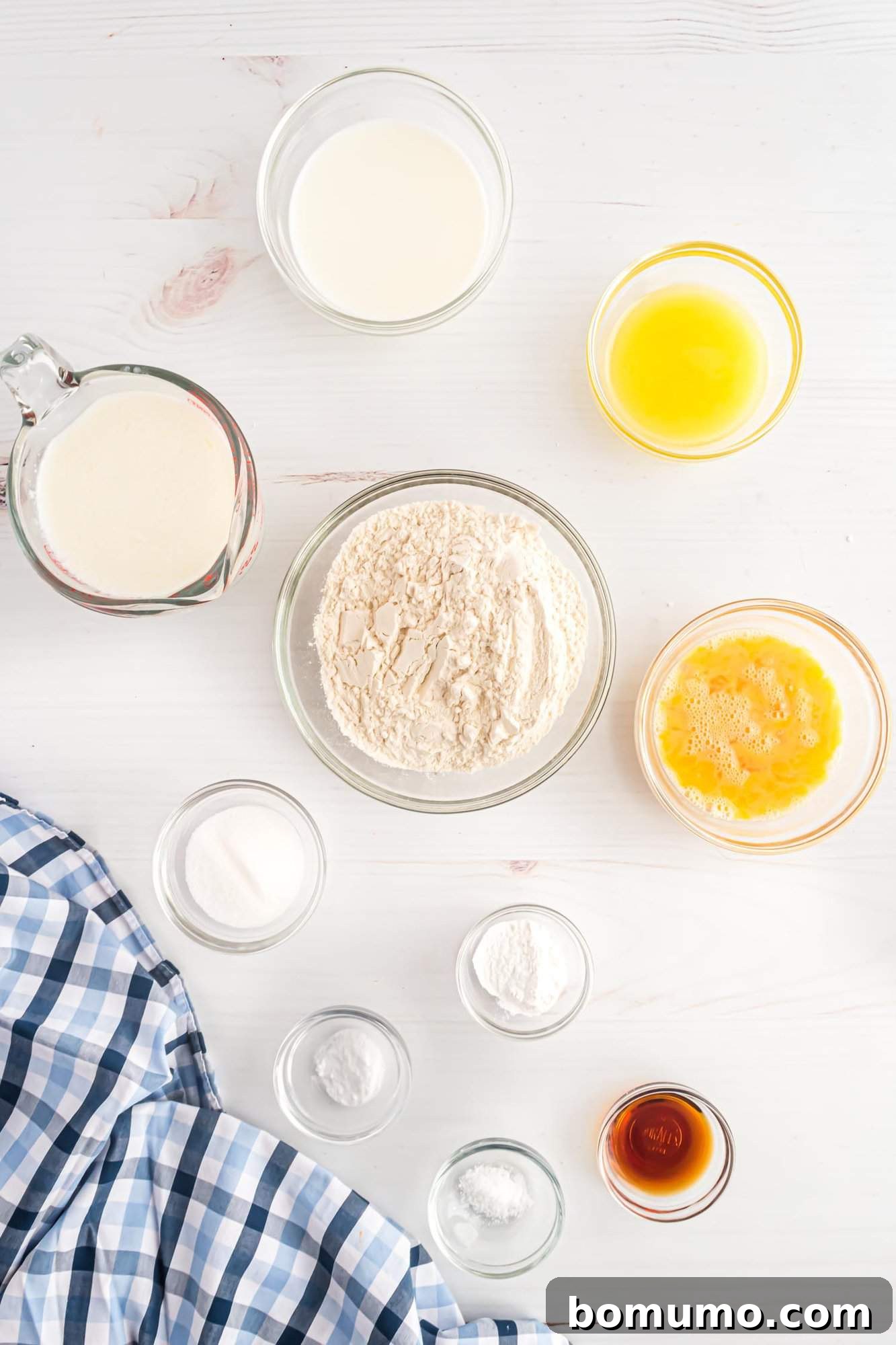 Various bowls containing flour, beaten eggs, melted butter, milk, vanilla extract, salt, baking powder, and sugar on a white surface with a checkered cloth in the corner.