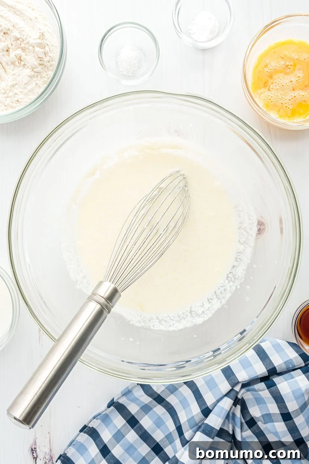 A whisk in a mixing bowl with wet ingredients, surrounded by flour, beaten eggs, and baking powder in small bowls. A blue and white checkered cloth is nearby.