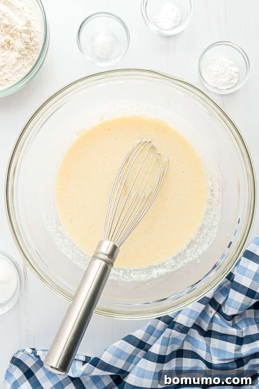 A whisk in a glass bowl containing wet ingredients. Surrounding the bowl are small glass bowls with various ingredients. A blue and white checkered cloth is partially visible.