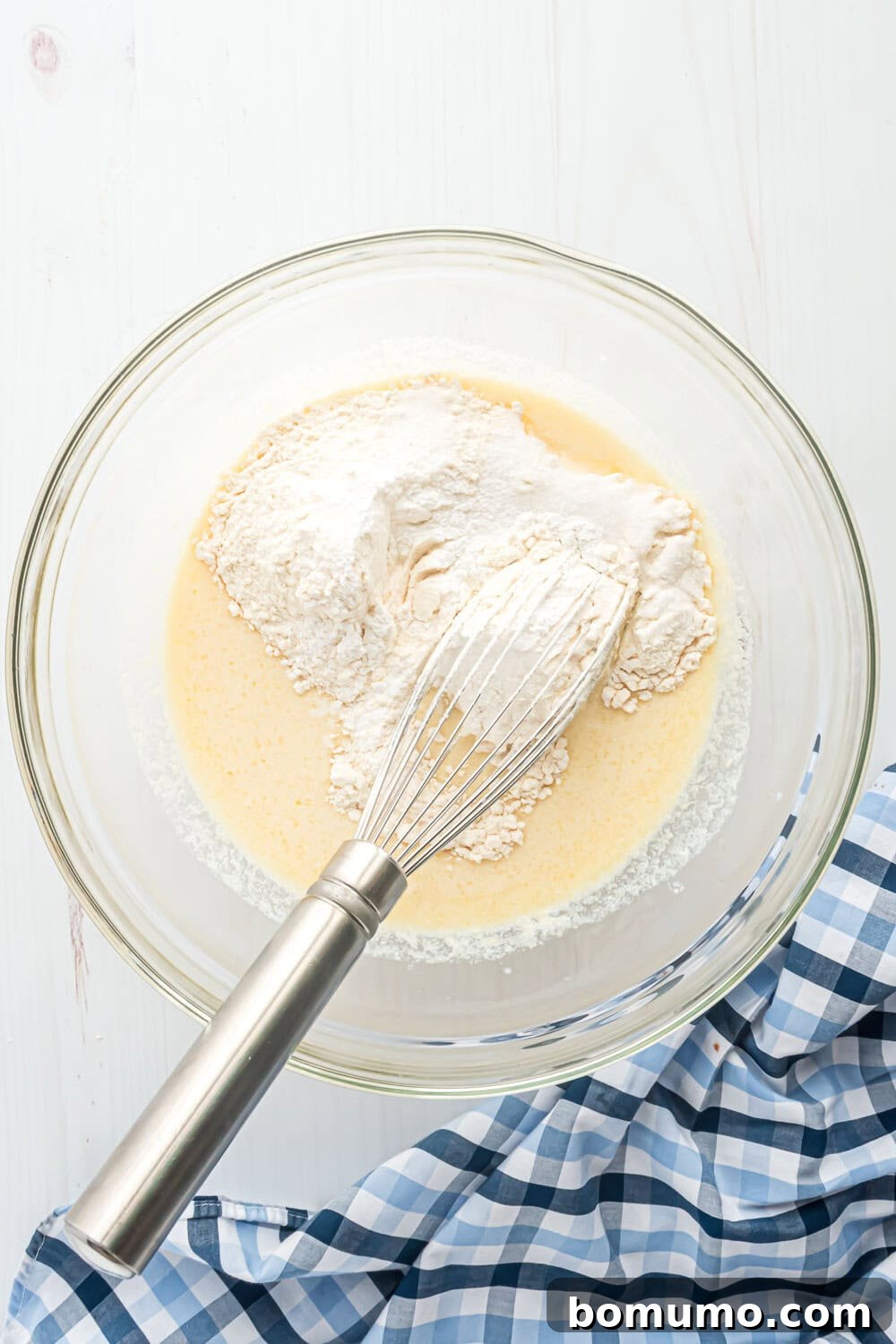 A whisk rests in a glass bowl containing flour, and wet ingredients, with a checkered cloth nearby.