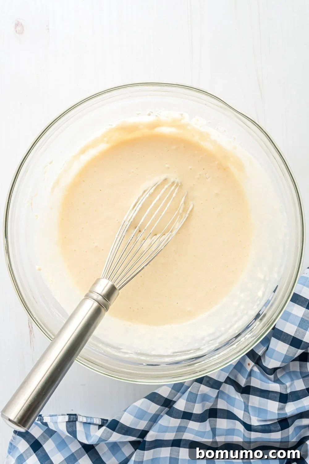 A whisk rests in a bowl of creamy batter on a white surface. A blue and white checkered cloth is partially visible beside the bowl.