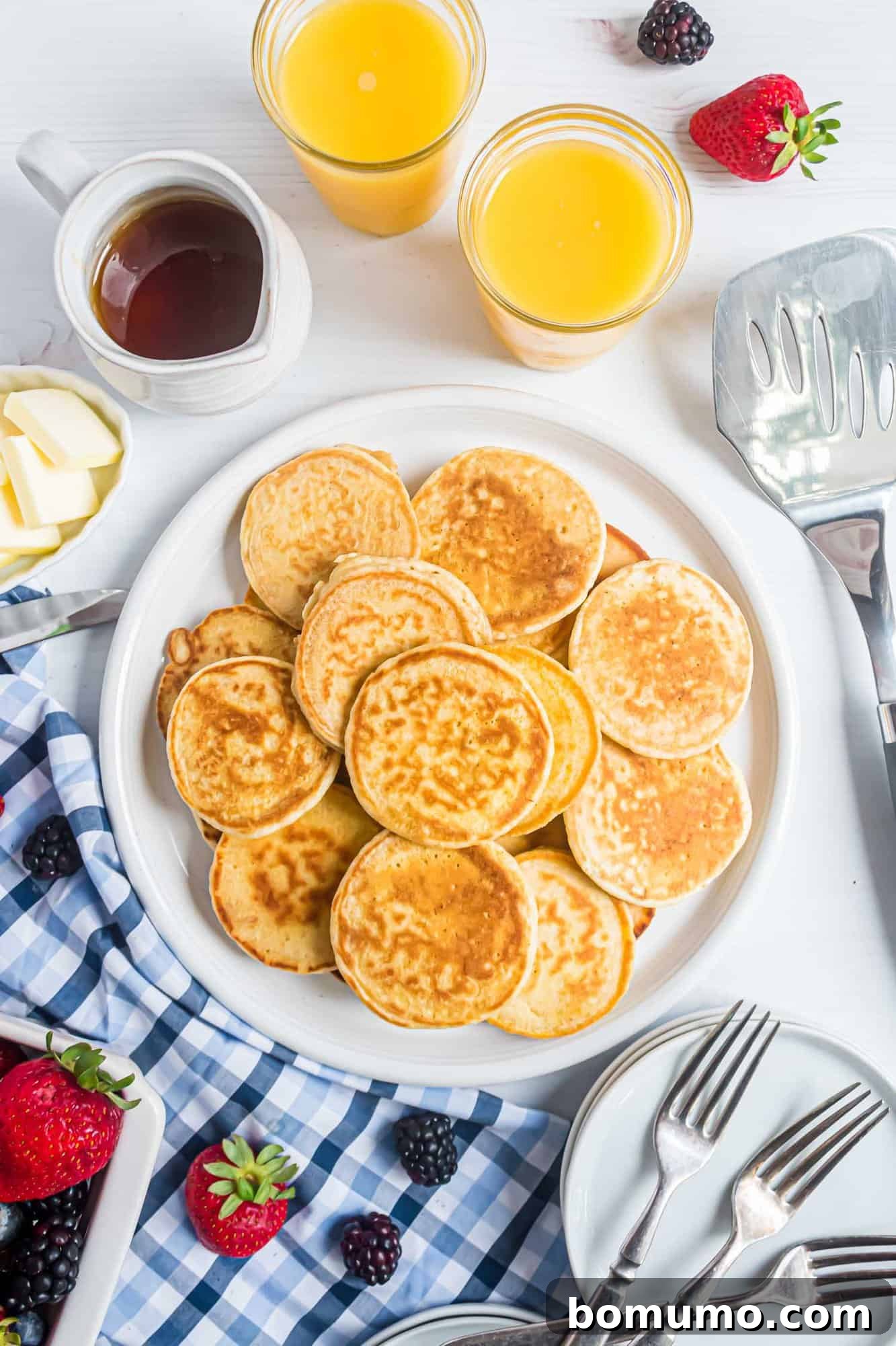 A plate of pancakes with butter, syrup, and two glasses of orange juice. Nearby are strawberries, blackberries, a spatula, and a blue checkered cloth with utensils.