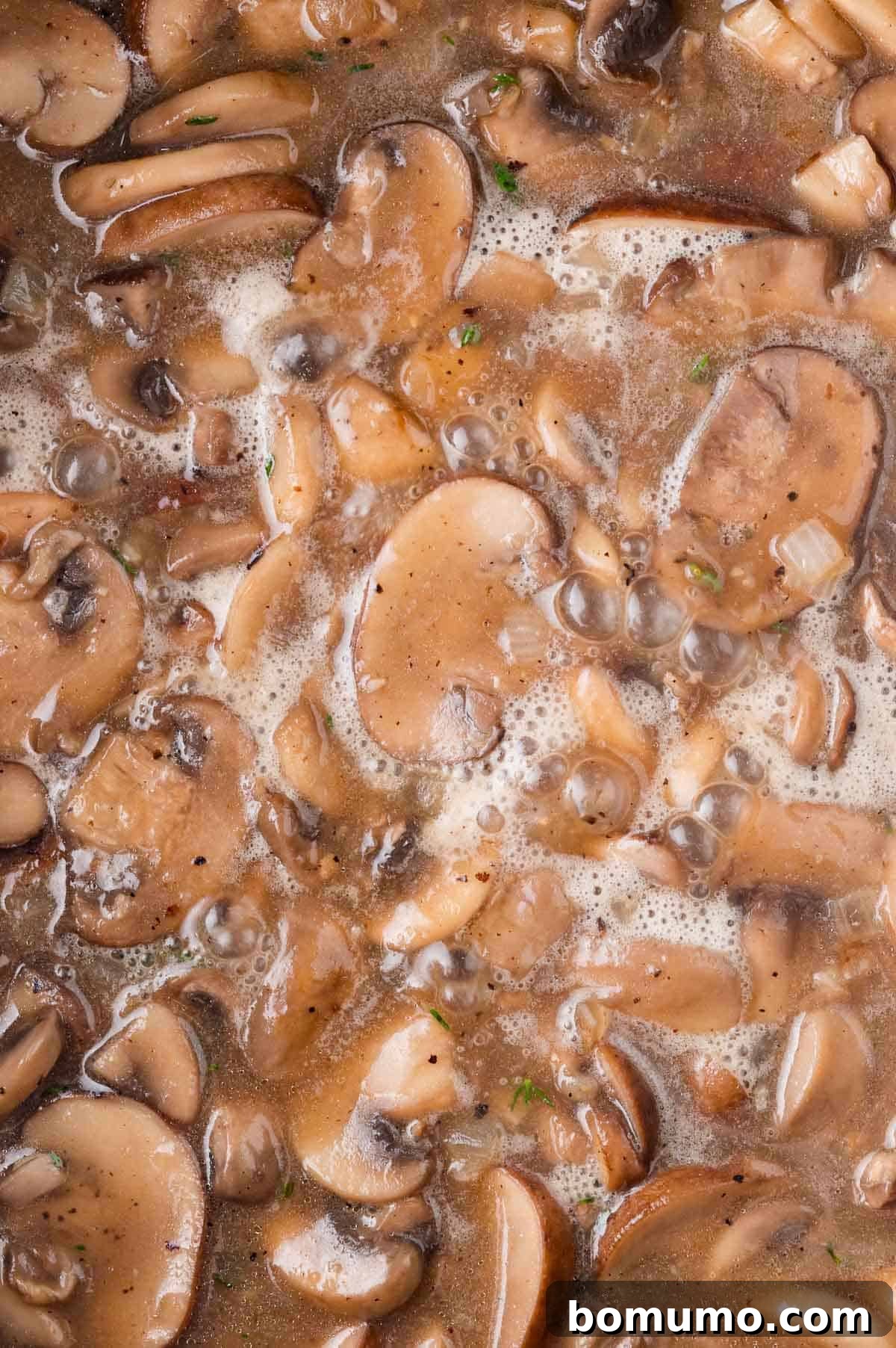 Close-up of sliced mushrooms cooking in a bubbling brown broth with visible herbs and seasonings.