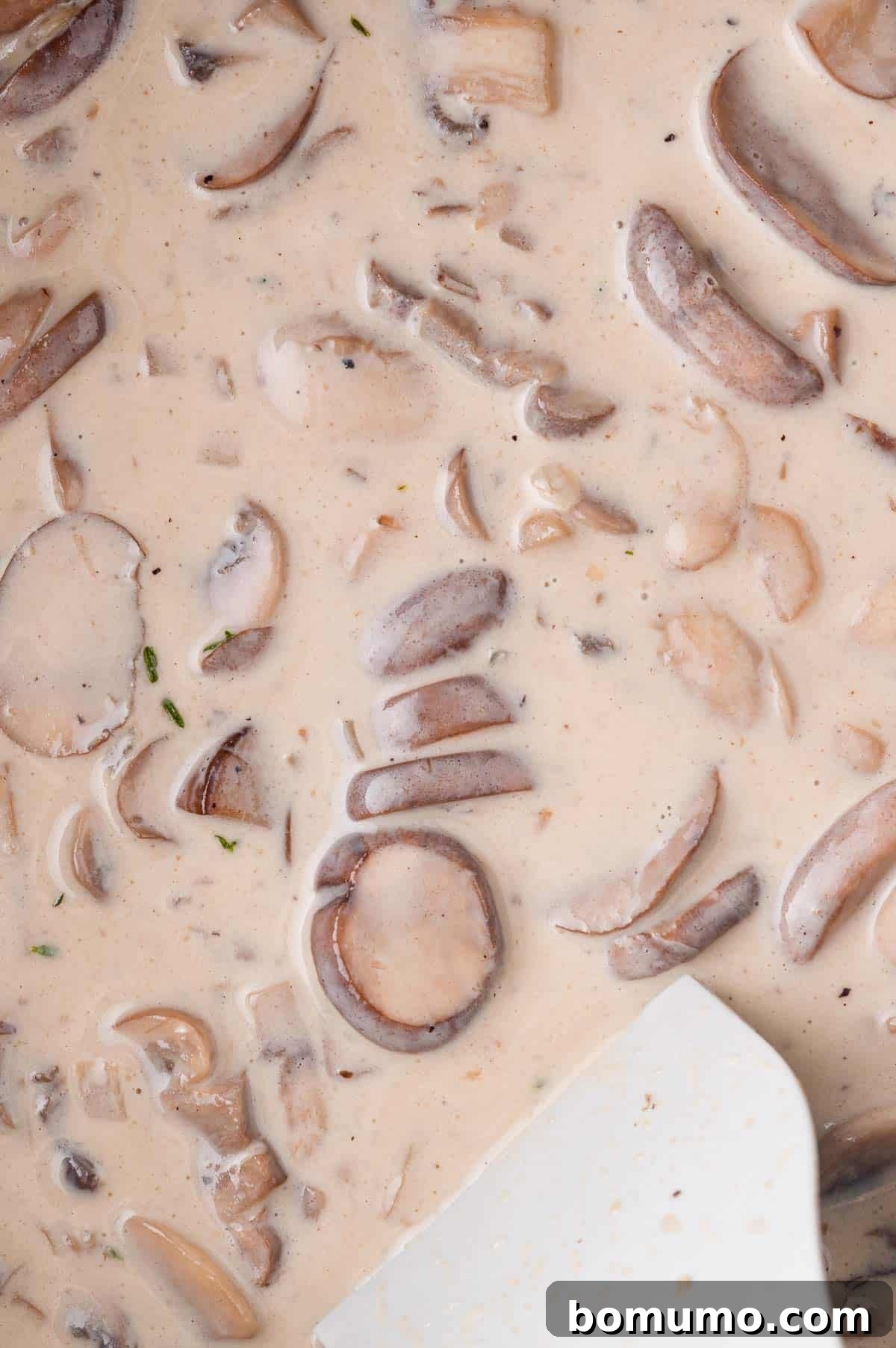 Creamy mushroom sauce with sliced mushrooms and herbs, partially mixed with a white spatula.