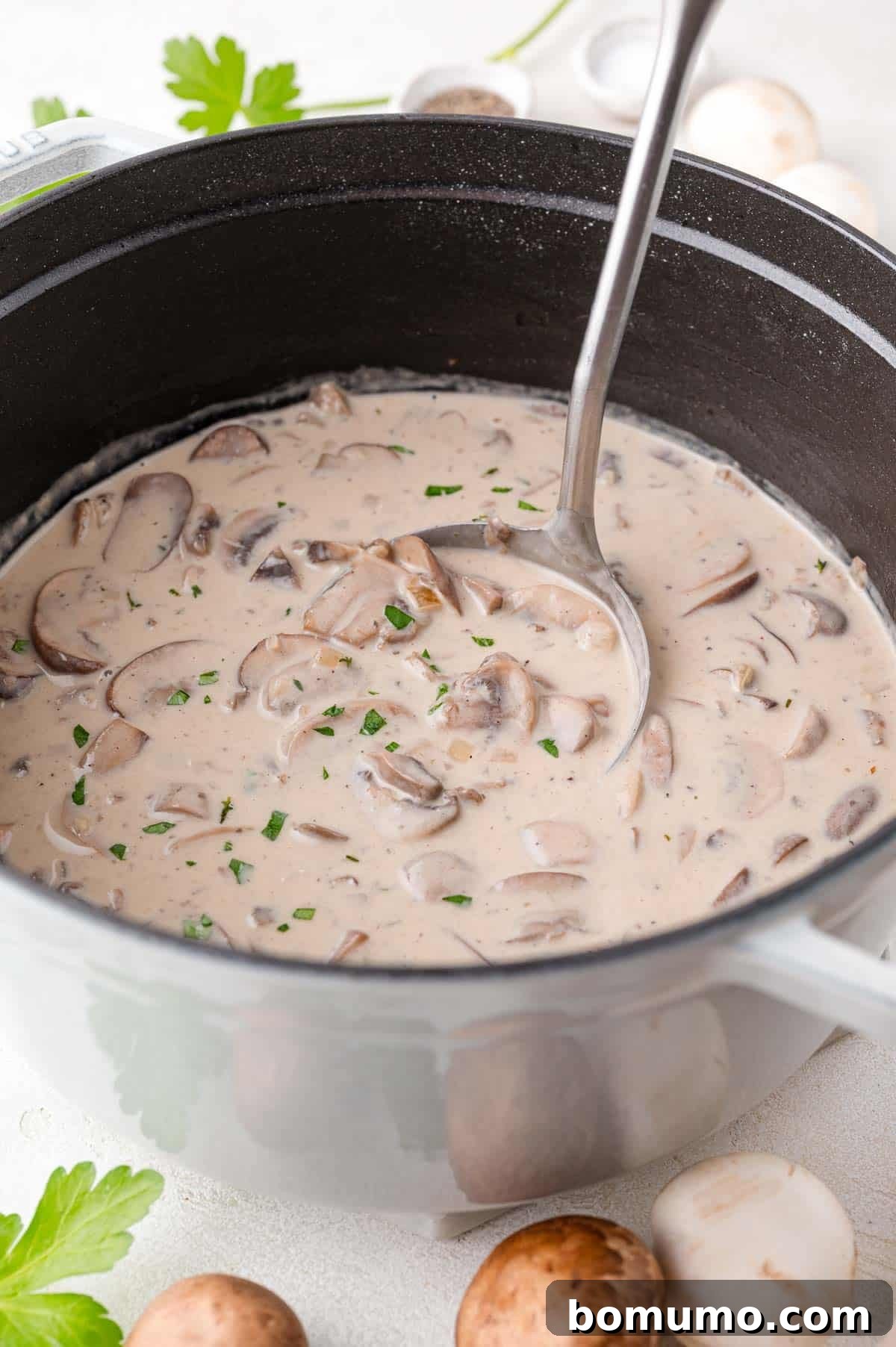 A pot of creamy mushroom soup with sliced mushrooms and chopped herbs, with a ladle.