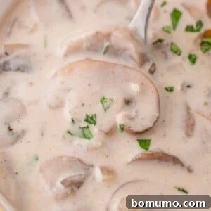 Close-up of creamy mushroom soup with sliced mushrooms and a garnish of chopped herbs, with a spoon partially visible.