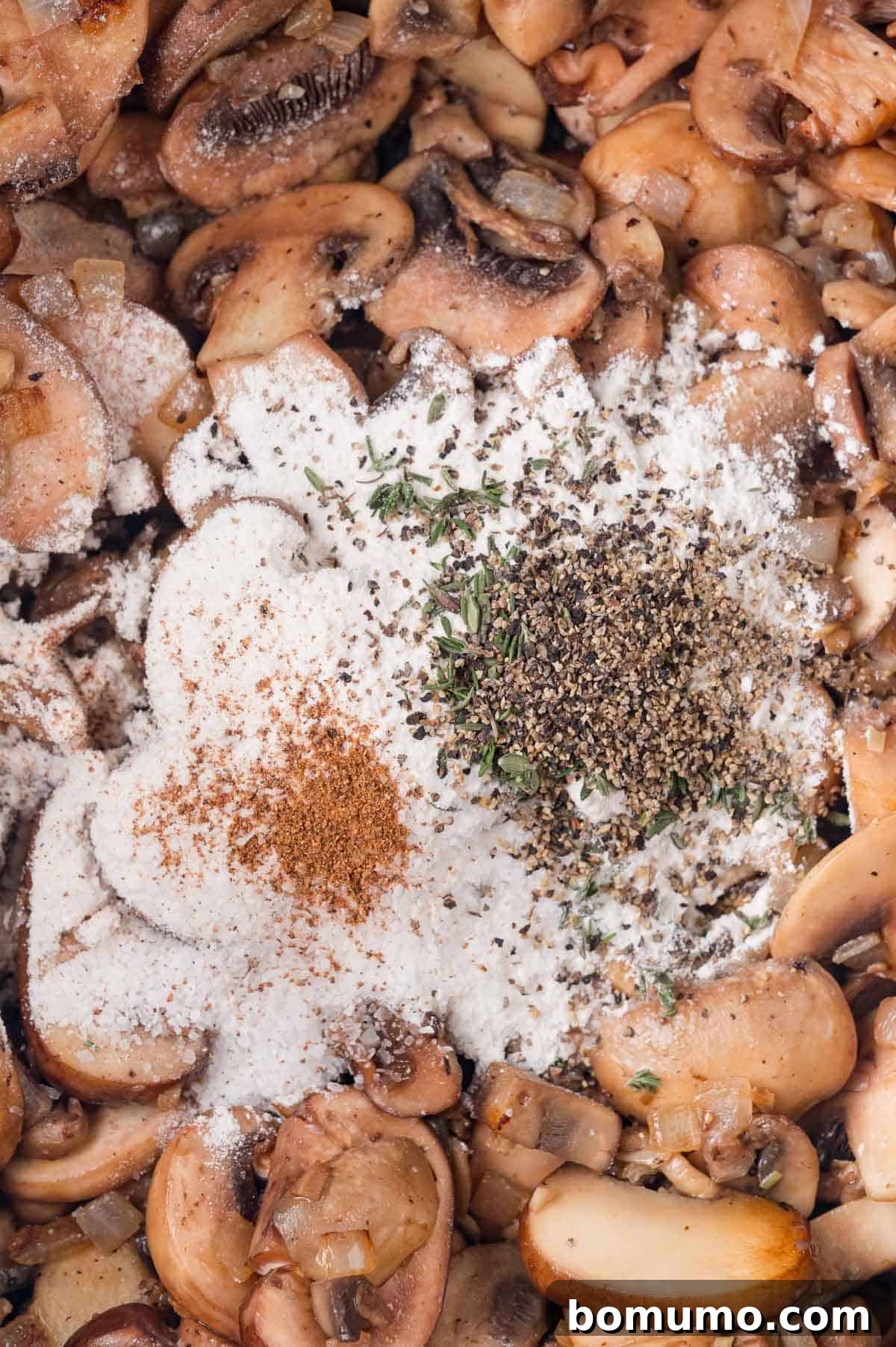 Close-up of sliced mushrooms with a dusting of flour, spices, and herbs, including black pepper and thyme, in a pan.