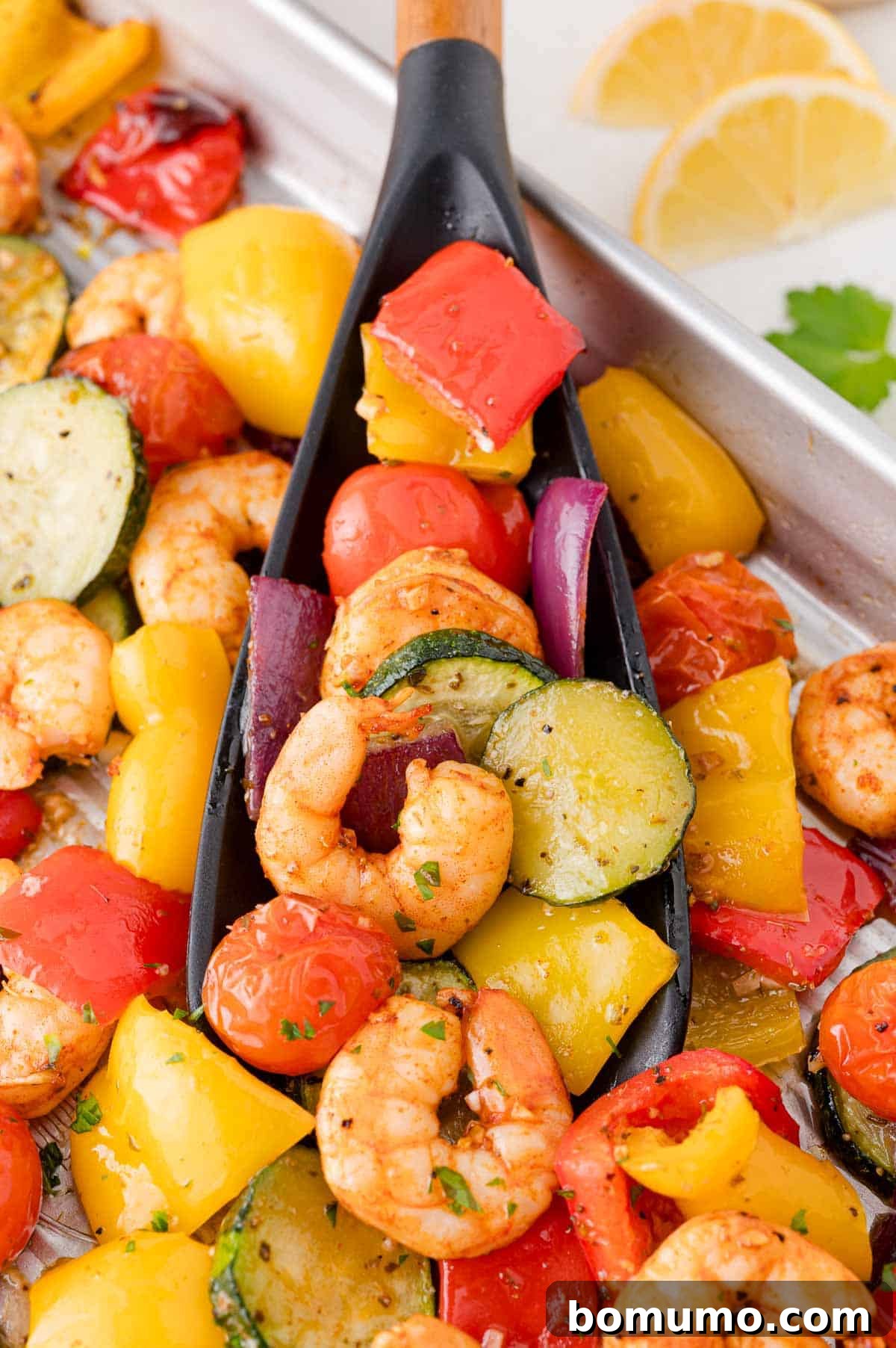 Close-up of a black spoon scooping mixed roasted shrimp and vegetables, including zucchini, cherry tomatoes, red onion, and bell peppers, from a metal tray. Lemon slices in the background.